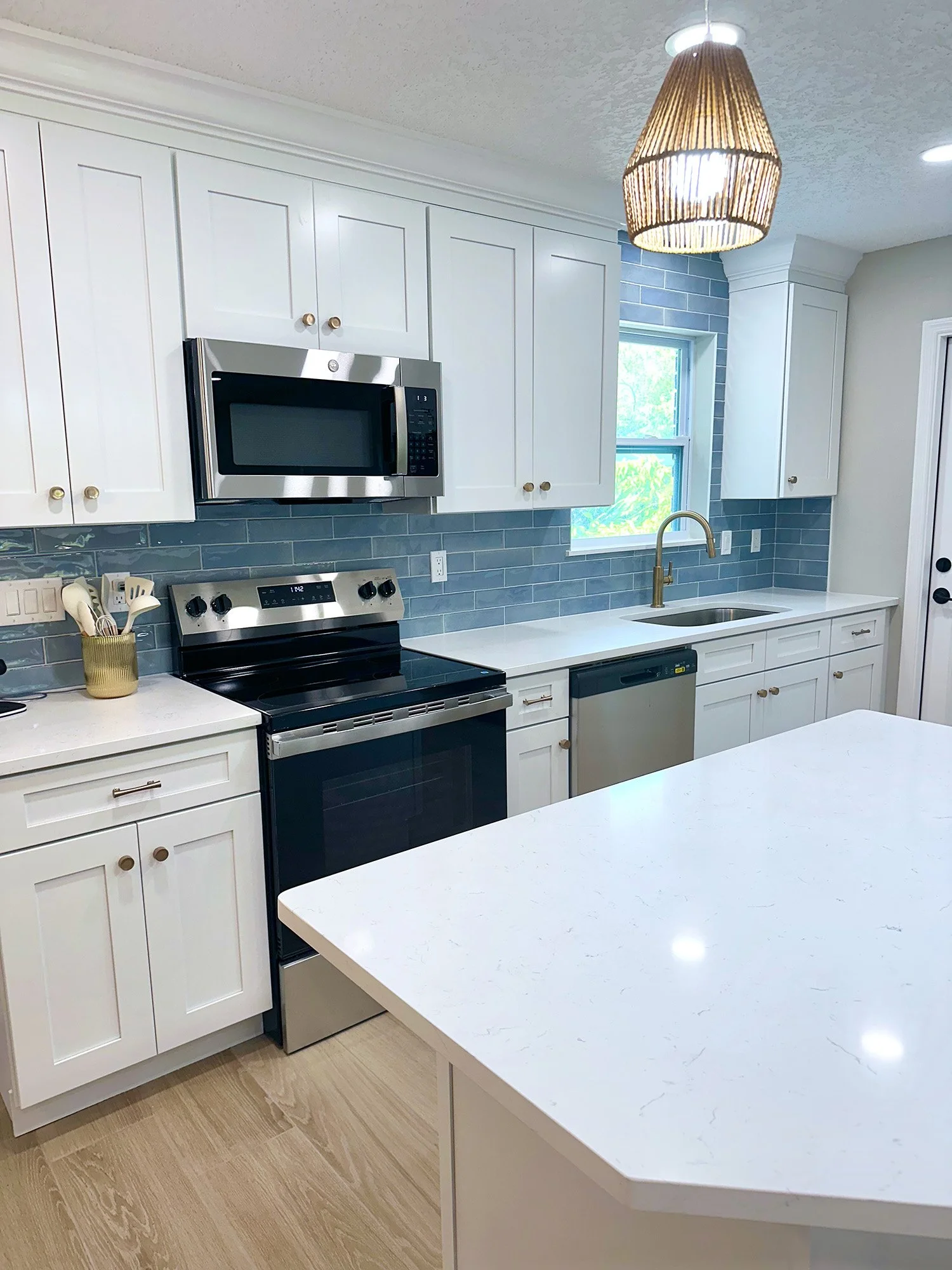 A modern kitchen with white cabinets, a stainless steel microwave, stove, and dishwasher, a white countertop, a gold faucet, a window with blue tiles around it, and a wicker pendant light.
