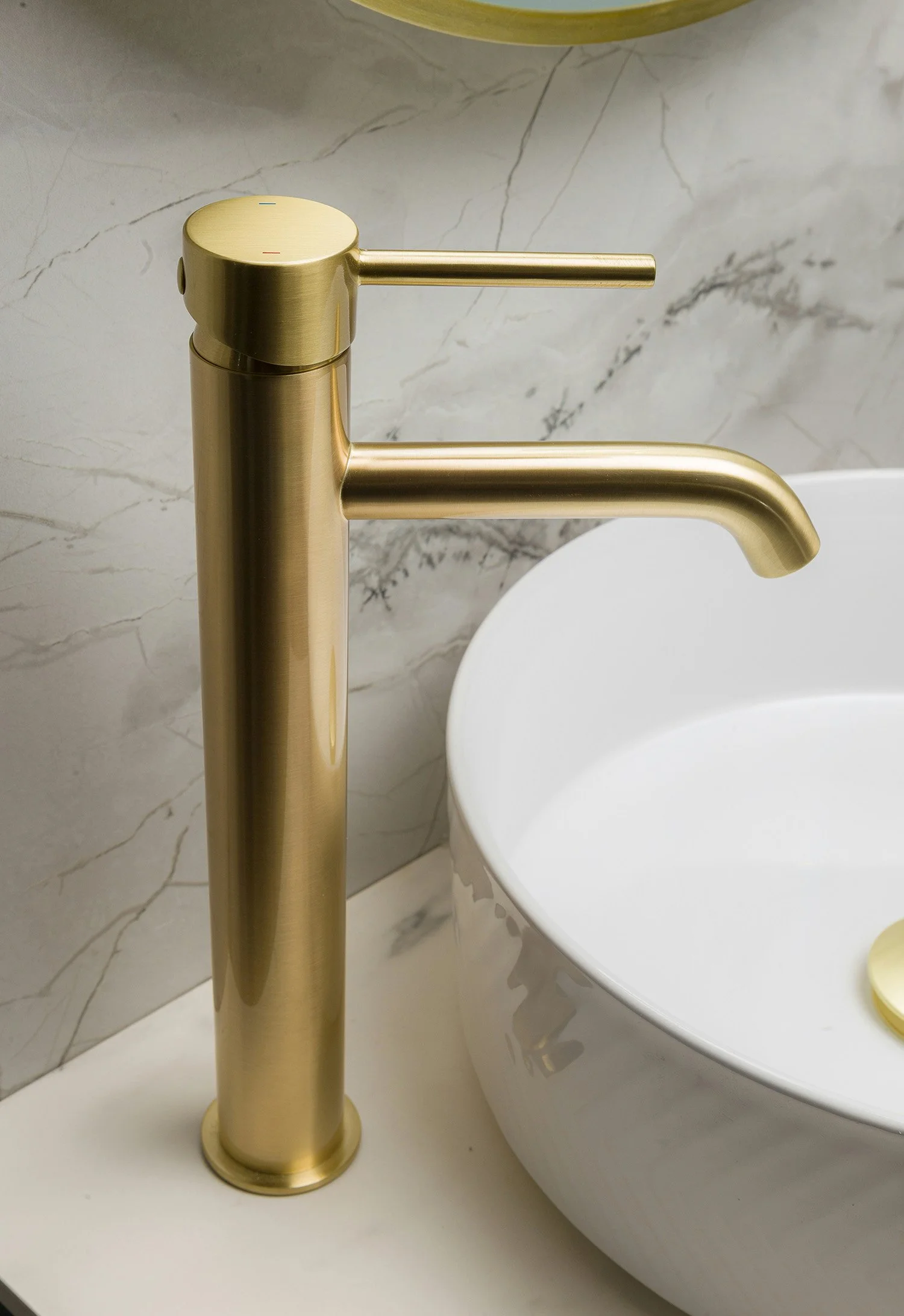 Close-up of a modern, gold-colored bathroom faucet next to a white oval sink with marble wall background.