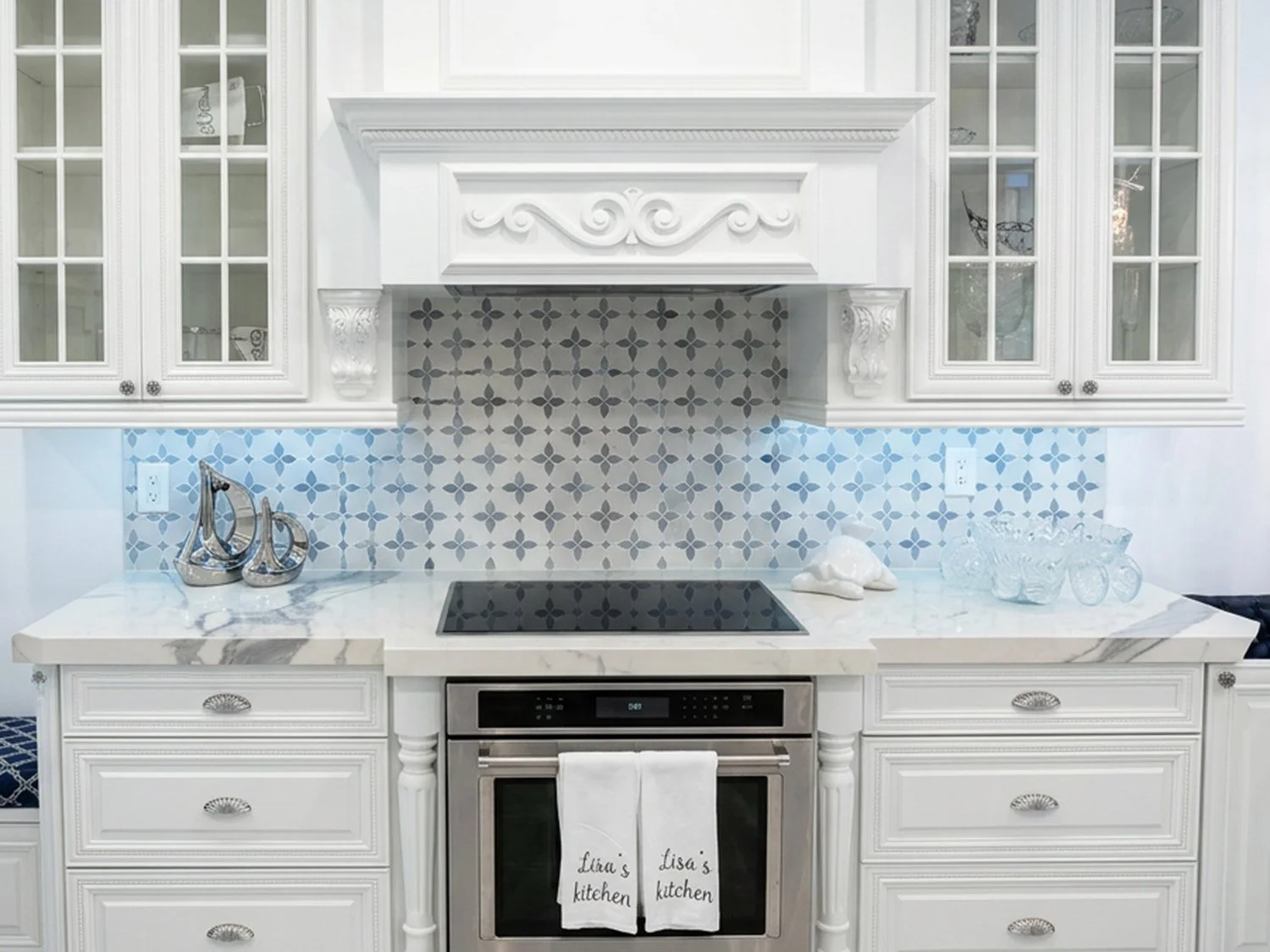 White kitchen with marble countertops, patterned backsplash, and decorated with silver sailboats and clear glass bowls. Stainless steel oven and stovetop with white towels hanging. Cabinets have glass doors with decorative molding.