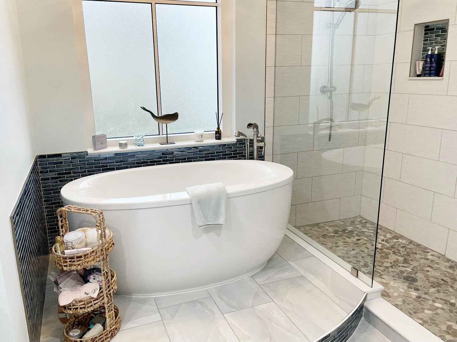 Modern bathroom with a freestanding white bathtub, a glass-enclosed shower, a window, and decorative items on the windowsill, including candles and a sculpture. A wicker basket filled with toiletries sits next to the bathtub.