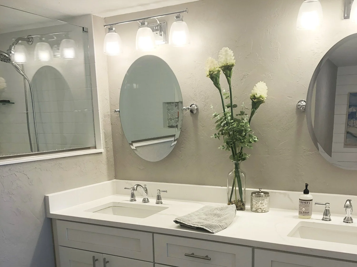 Bathroom vanity with two sinks, mirrors, a vase with white flowers, and bathroom items on the countertop.