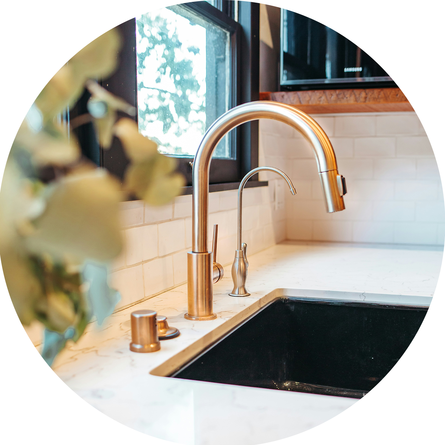 Close-up of a kitchen sink with a gold-colored faucet and marble countertops, a window with a view of trees outside