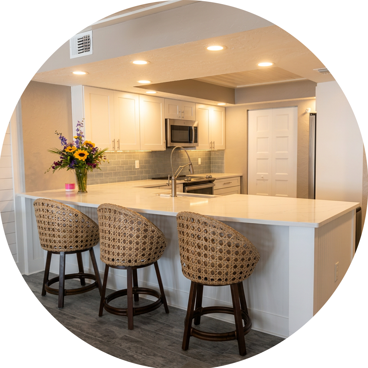 Modern kitchen with white cabinets, a marble island with three rattan barstools, a bouquet of sunflowers and purple flowers in a glass vase, and recessed ceiling lights.