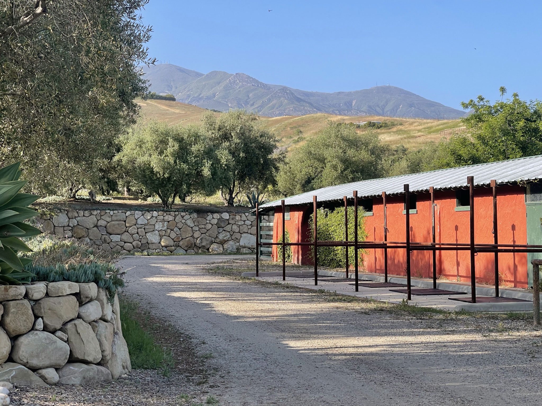 A landscape view of a quiet outdoor area with a stone wall, a red building with a metal roof, surrounding trees, and mountains in the background under a clear blue sky.
