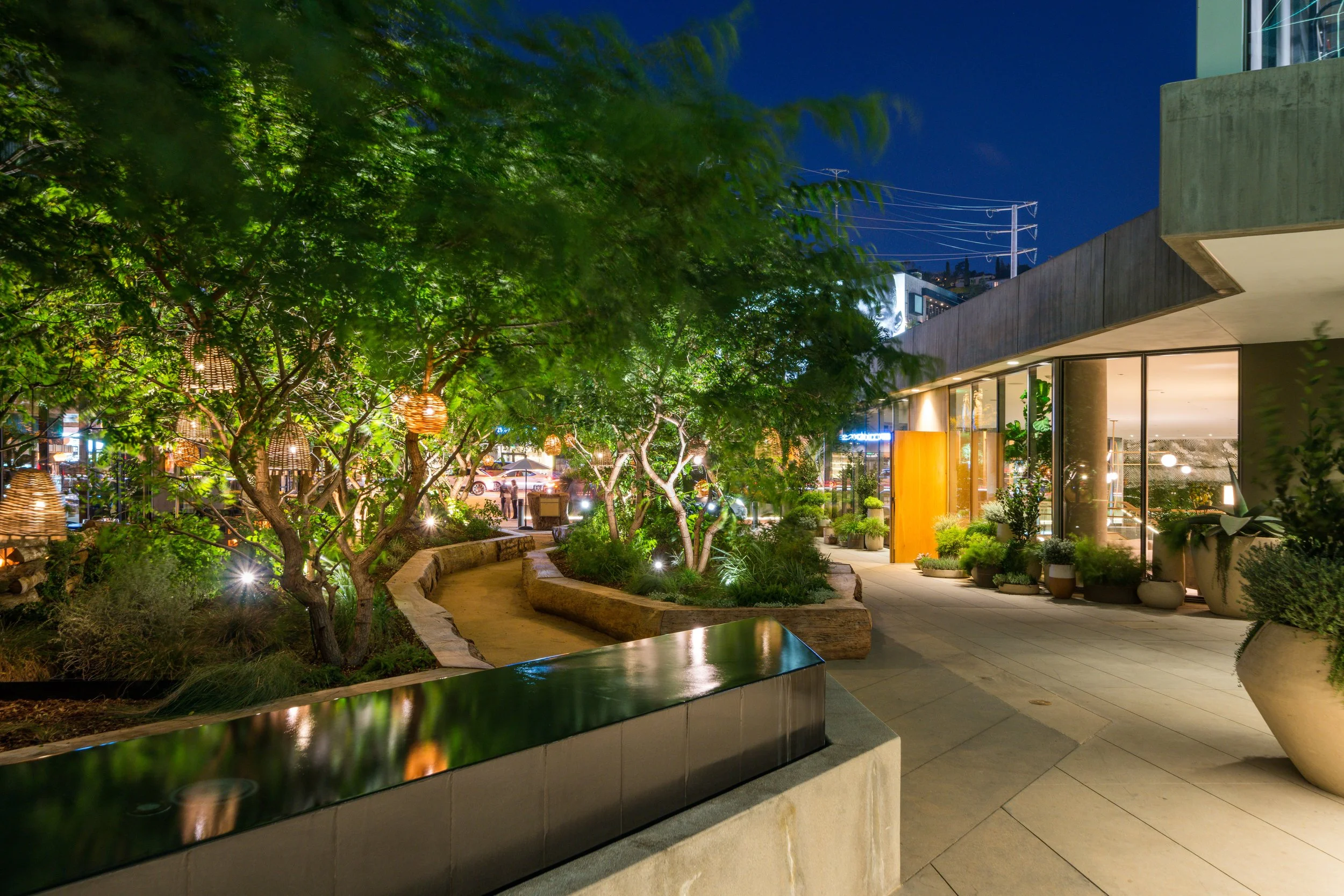 Nighttime scene of an outdoor urban area with a modern building, illuminated trees with hanging lanterns, and people walking along a pathway.