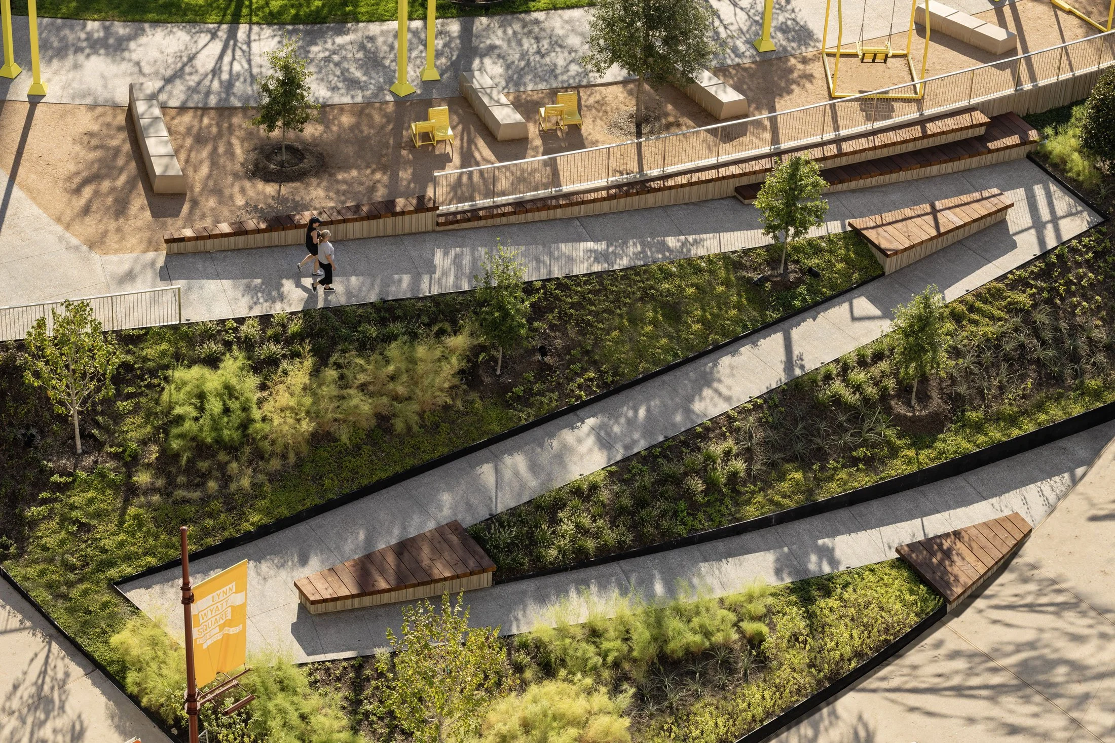 An aerial view of a modern urban park with paved walkways, green trees, benches, and landscaped areas. Two people are walking along a pathway near the bottom left.