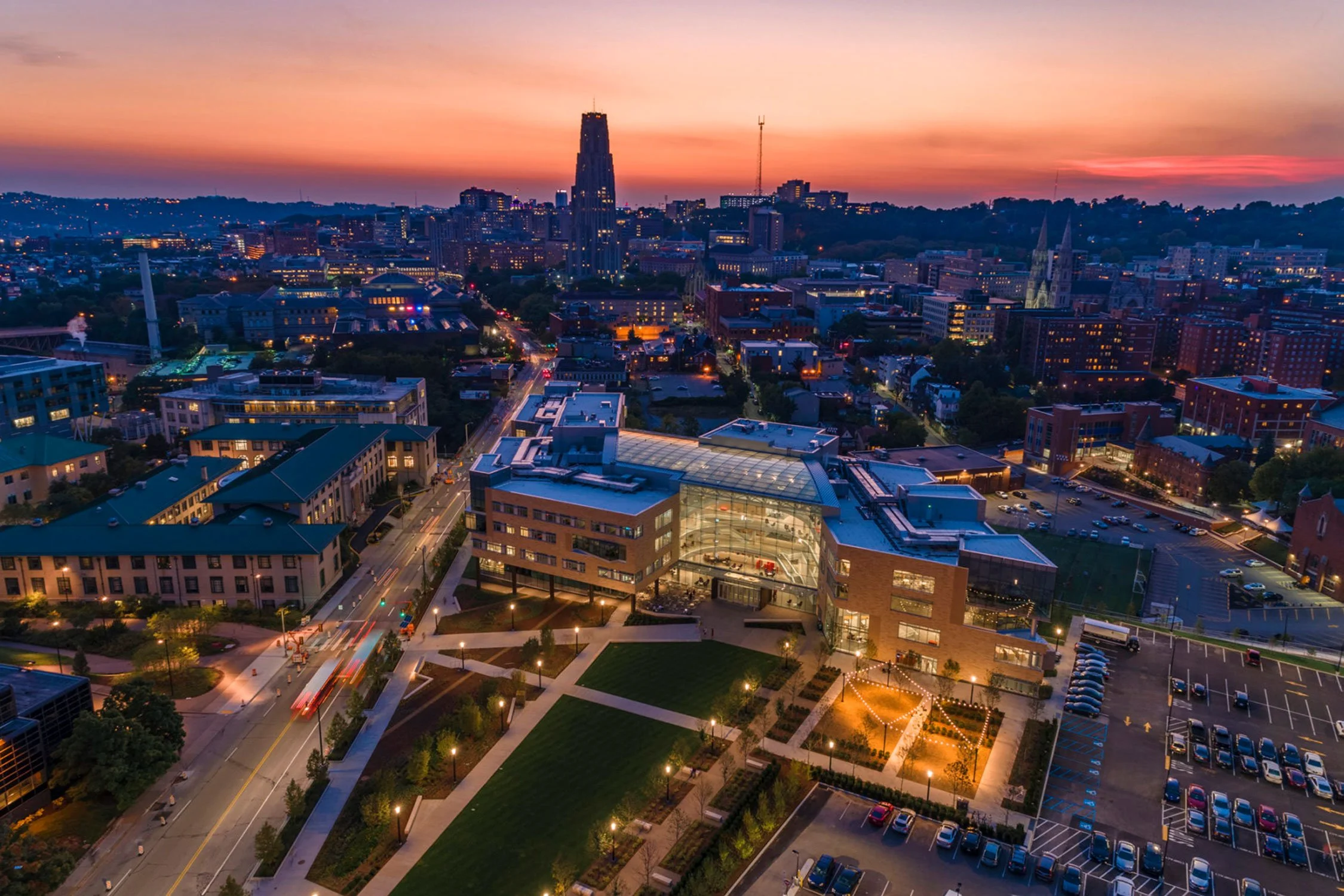 City skyline at sunset with buildings, streets, parking lots, and a park with green lawns and trees in downtown area.