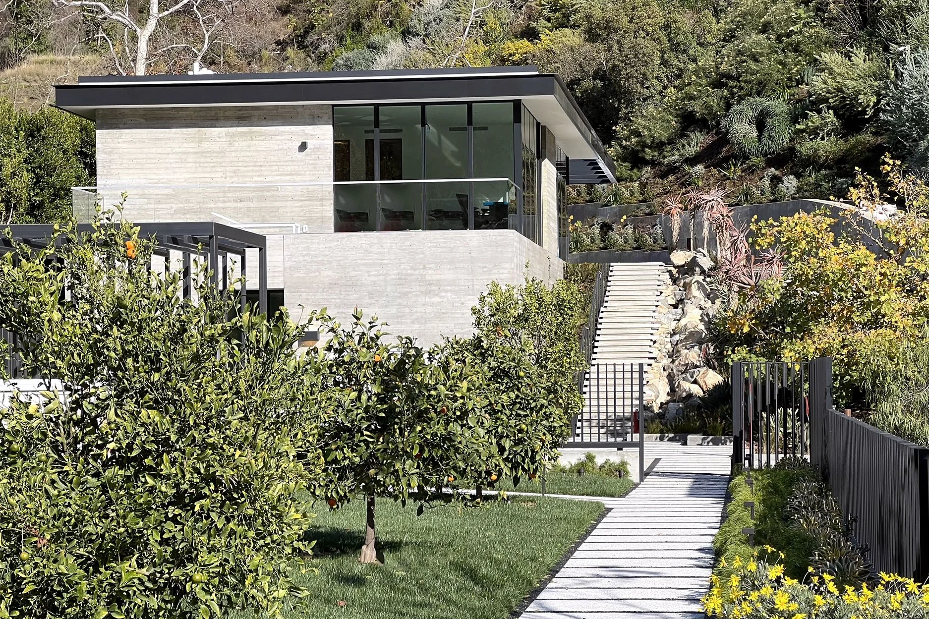 Modern house with large glass windows, surrounded by lush greenery, a stone staircase, and a garden with trees and shrubs.