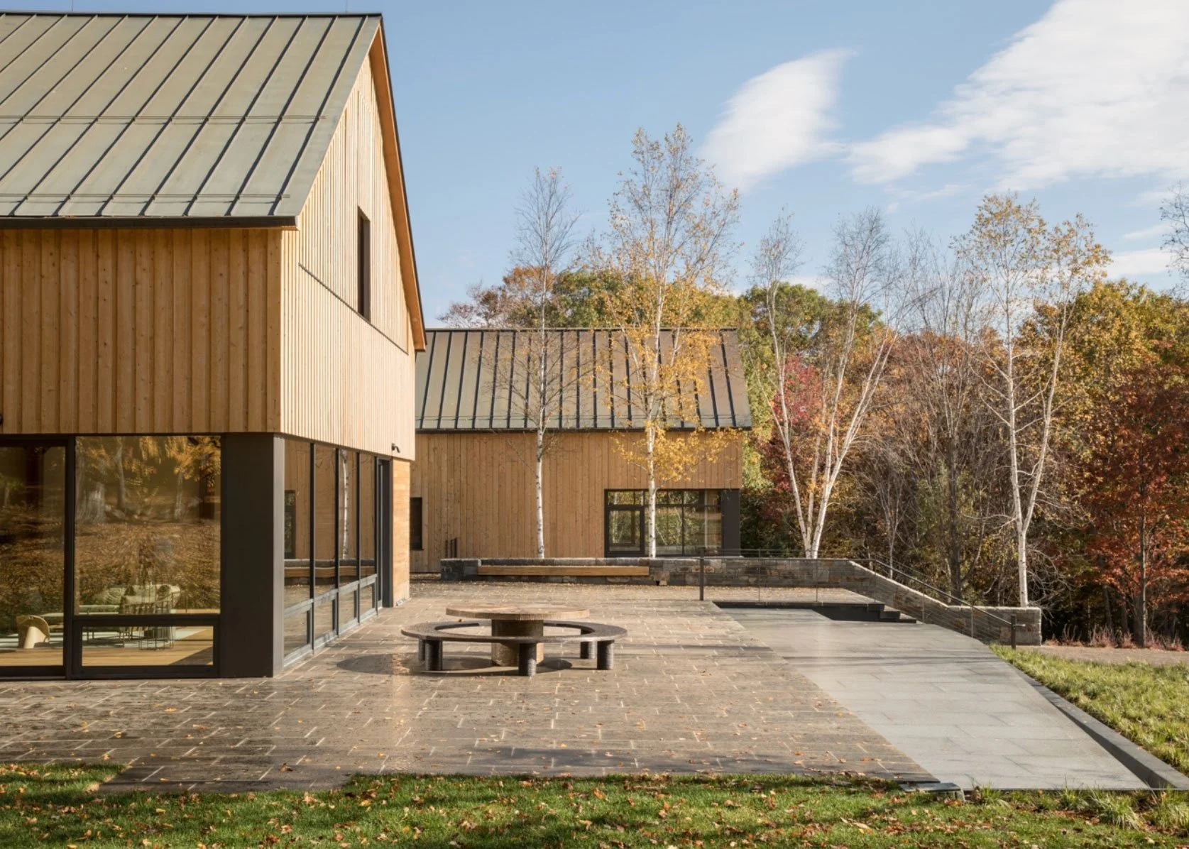 Modern wooden building with large glass windows, surrounded by fall-colored trees and an outdoor patio with a picnic table, in a natural setting.
