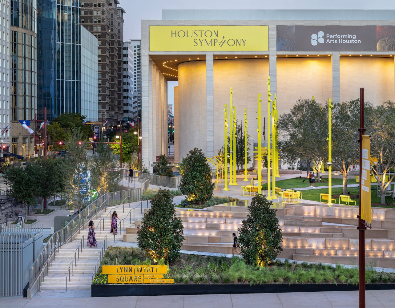 A city park in Houston, Texas, with modern yellow benches, bike racks, trees, and illuminated landscaped steps, surrounded by high-rise buildings and a sign indicating 'Lynn Wyatt Square at the Houston Symphony'.