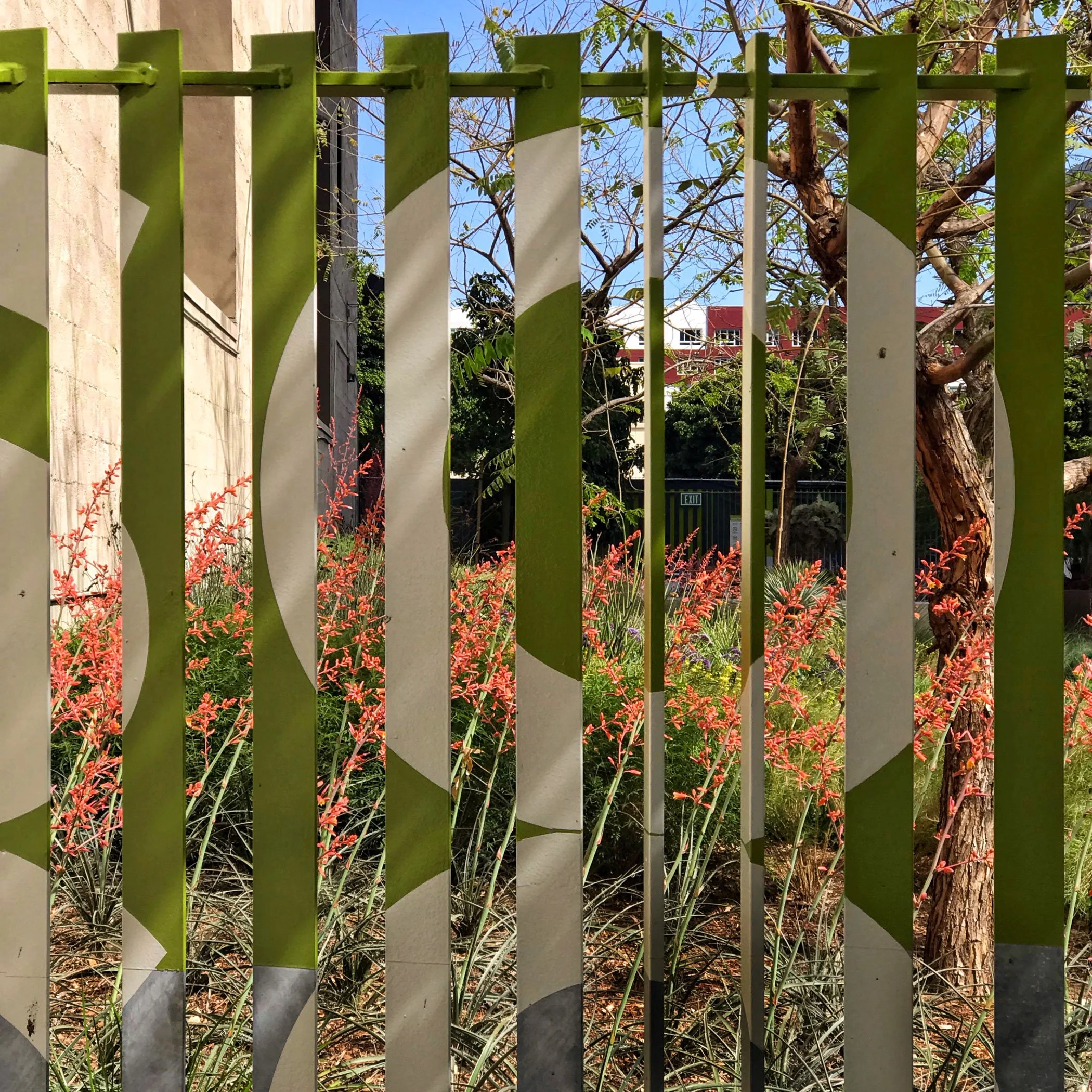 Green and white metal fence with plants, trees, and buildings in the background under a blue sky.
