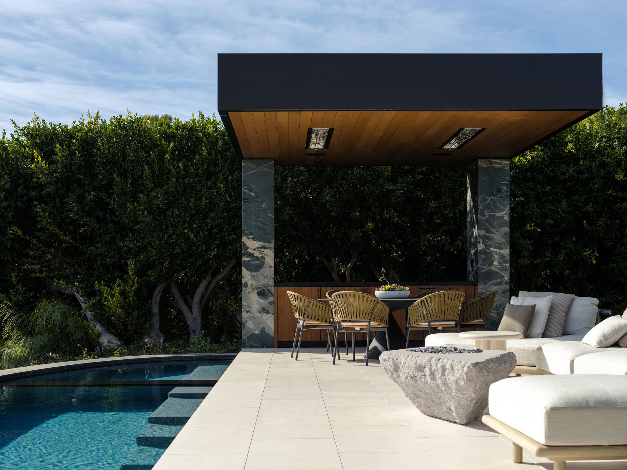 Modern backyard with a swimming pool, outdoor dining area with chairs, a seating area with pillows, a fire pit, and lush green hedges and trees in the background.
