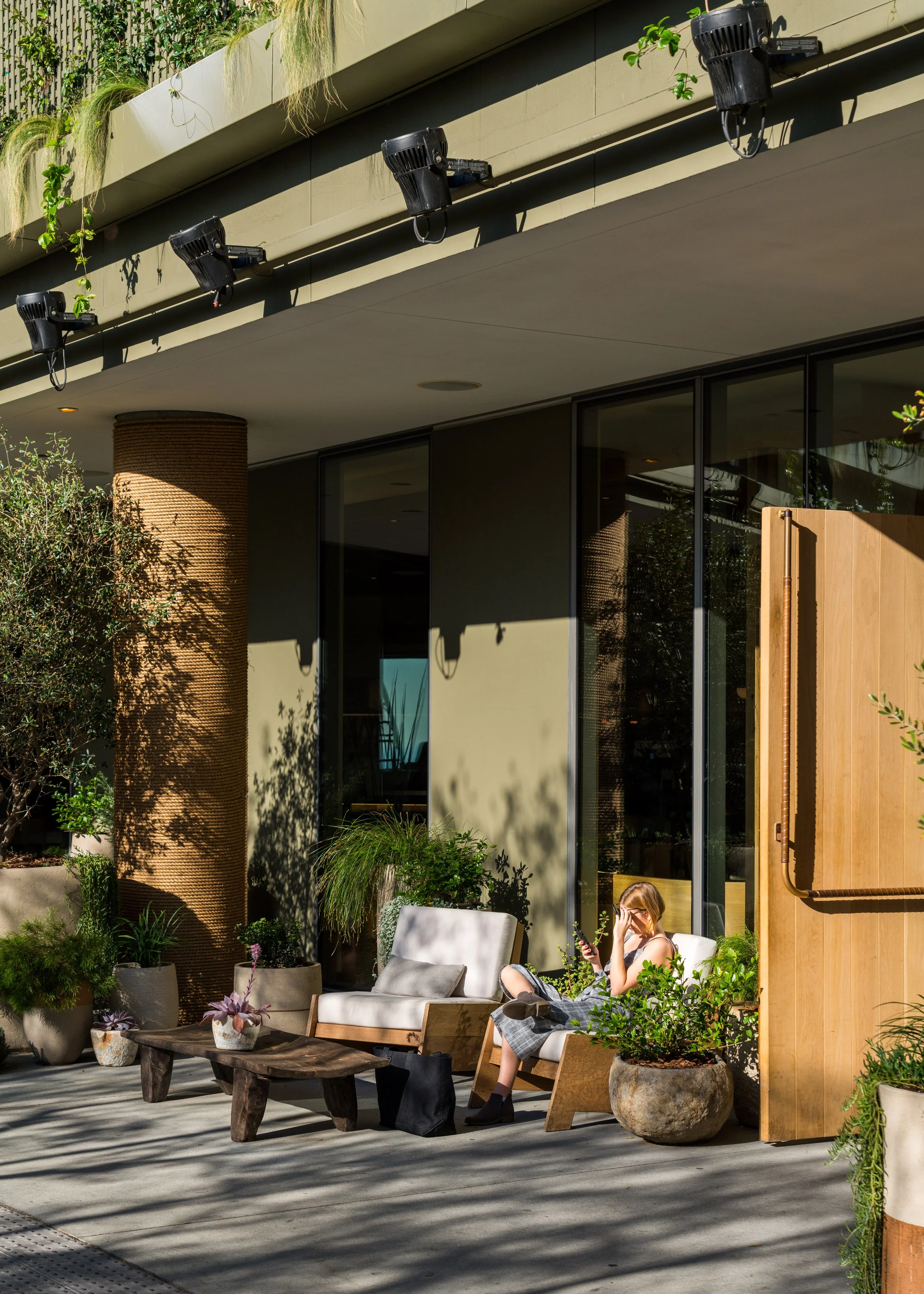 An outdoor patio with potted plants, a woman sitting on a cushioned wooden lounge chair, and a woman with blonde hair sitting on a chair, using her phone, surrounded by plants and modern architecture.