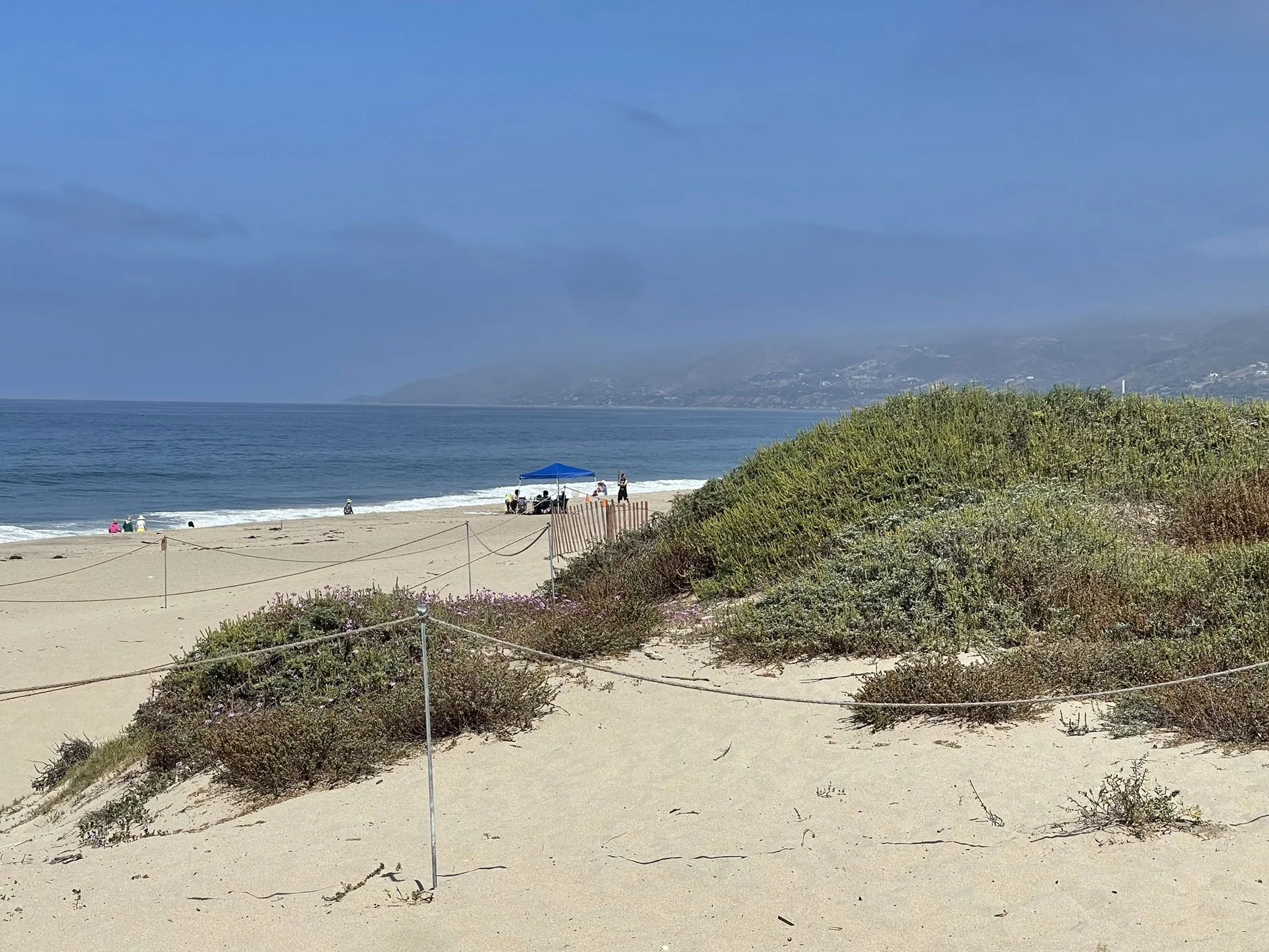 Beach with sandy dunes in the foreground, green bushes along a rope barrier, people sitting and standing under a blue canopy, the ocean with small waves in the middle ground, and hills with houses and smoke in the background under a blue sky with clo