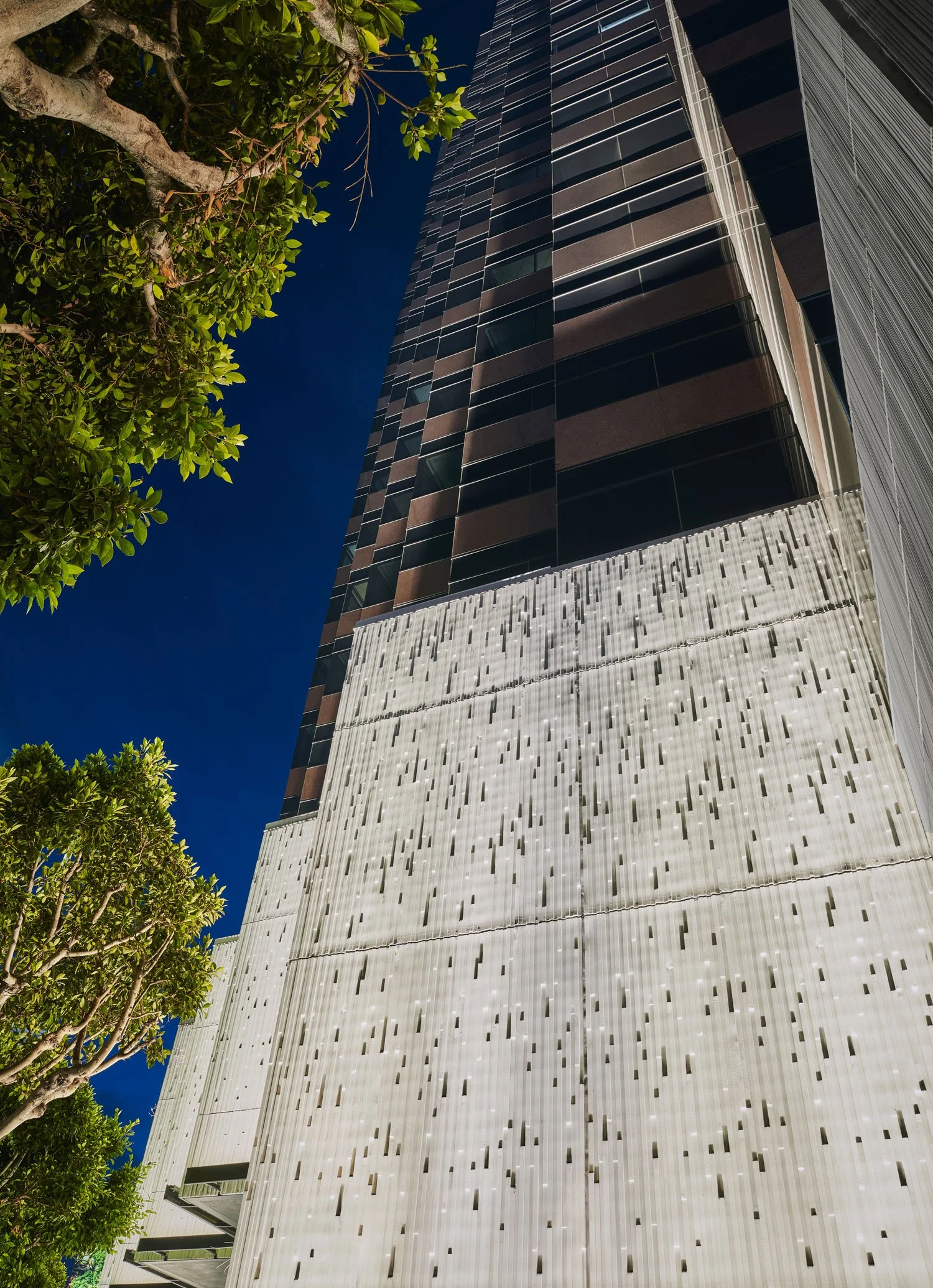 Low-angle view of a modern building with a textured white facade, black glass windows, and dark horizontal panels, with green trees in the foreground under a clear blue sky at night.