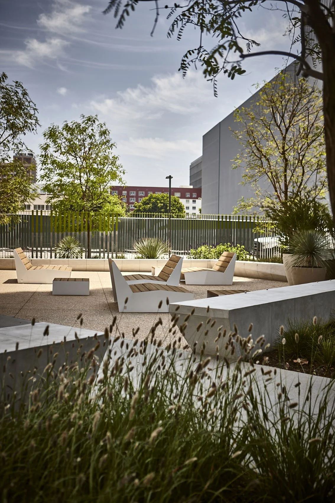 An outdoor patio with modern patio furniture, including four lounge chairs with wooden slats and white supports, set on a beige paved surface, surrounded by potted plants and trees, with a cityscape and a partly cloudy sky in the background.