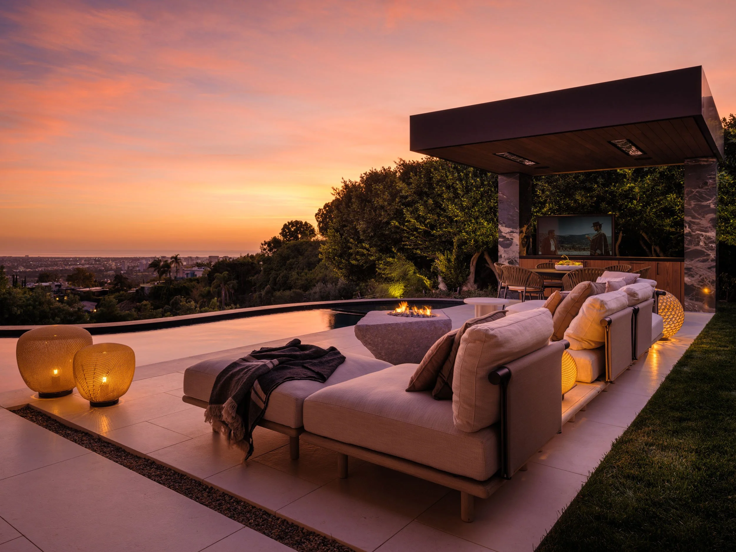 Luxury outdoor patio at sunset with modern sofa, throw blankets, and lanterns near a reflecting pool, overlooking a city view with trees and buildings.