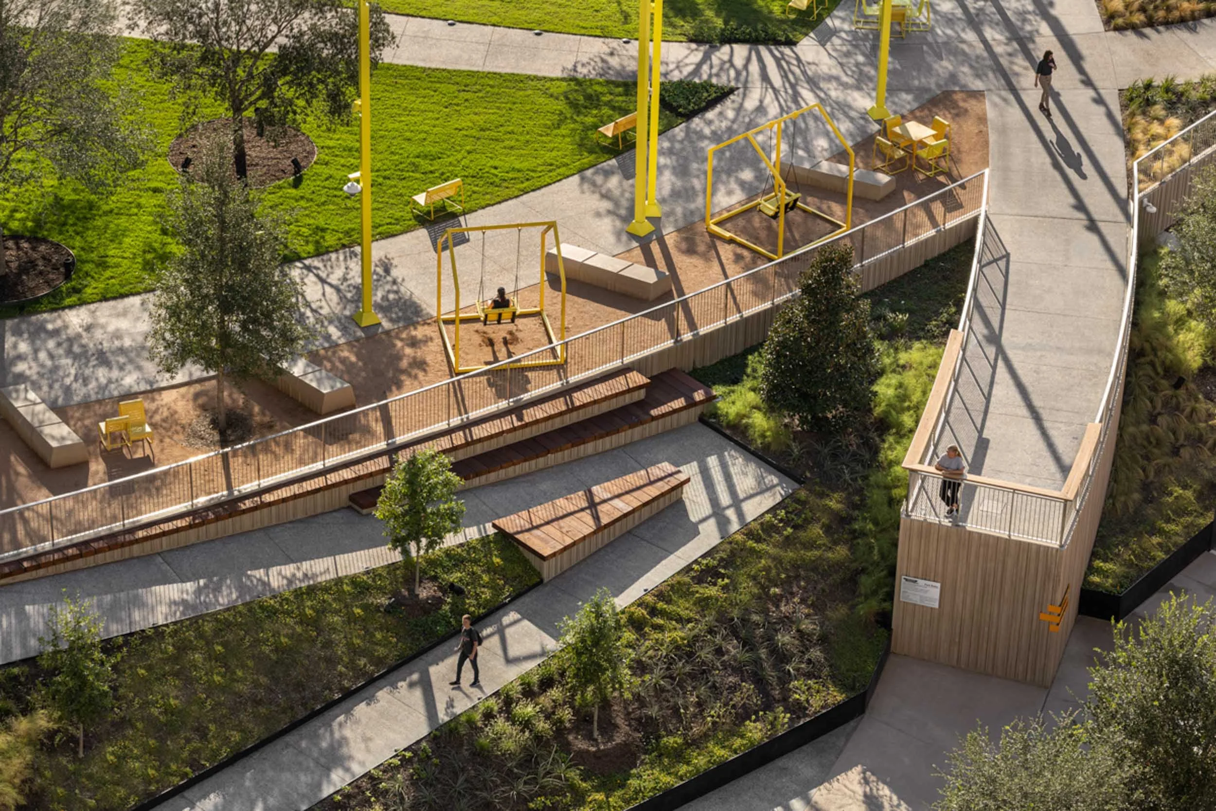 An aerial view of a park with green grass, trees, and pathways. There are yellow swings and seating areas along the walkways, with a few people walking and sitting.