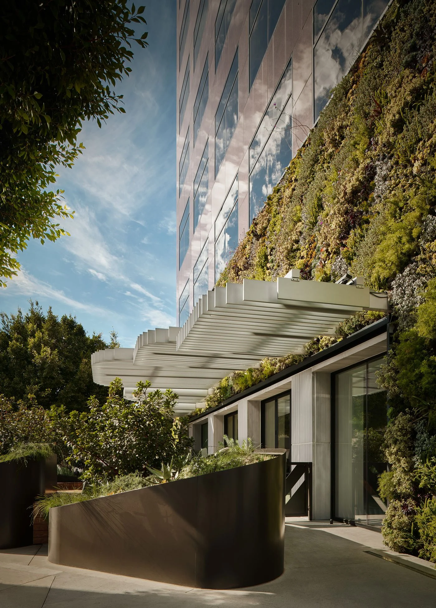 Modern building with glass windows reflecting the sky and clouds, surrounded by a lush garden with various plants and trees, and featuring white architectural elements.