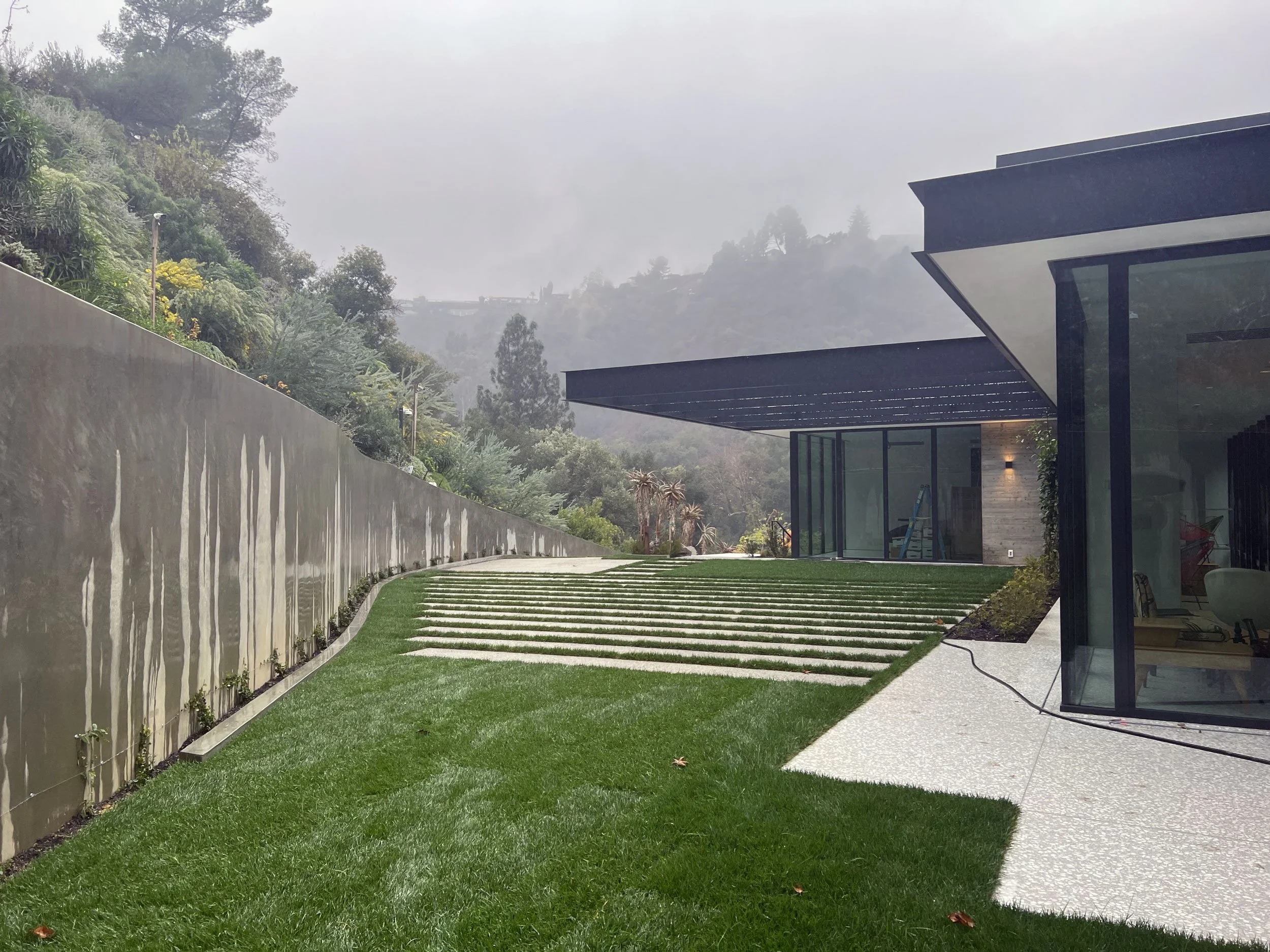 Modern house with large windows and black exterior, overlooking a lush green yard with grass and pathway, with misty mountains and trees in the background.