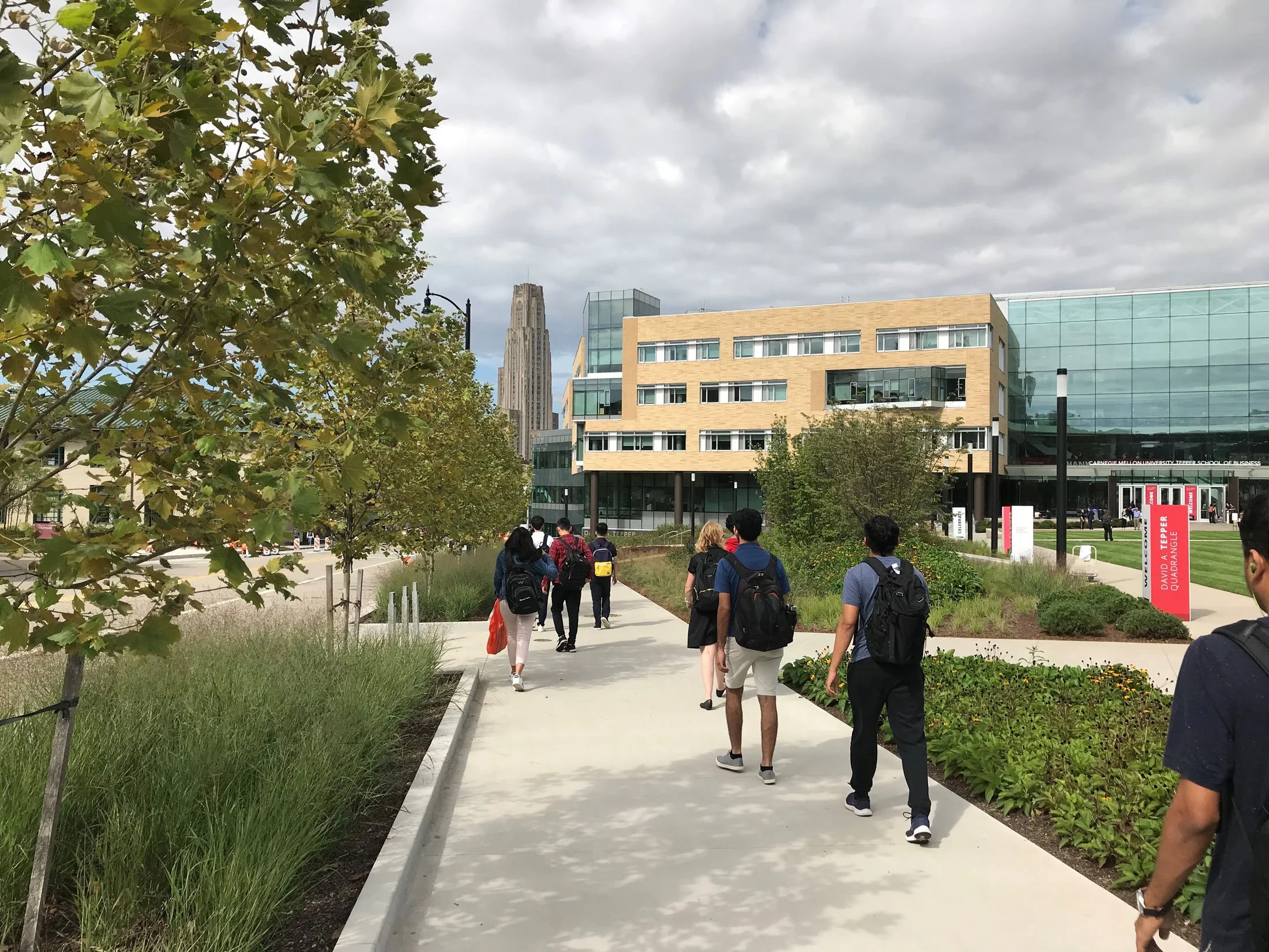 People walking on a sidewalk in Pittsburgh's Carnegie Mellon University's urban campus setting with modern buildings and trees, enhanced with streetscape improvements such as planters, plantings, adjacent seating, and ample access to and from campus.