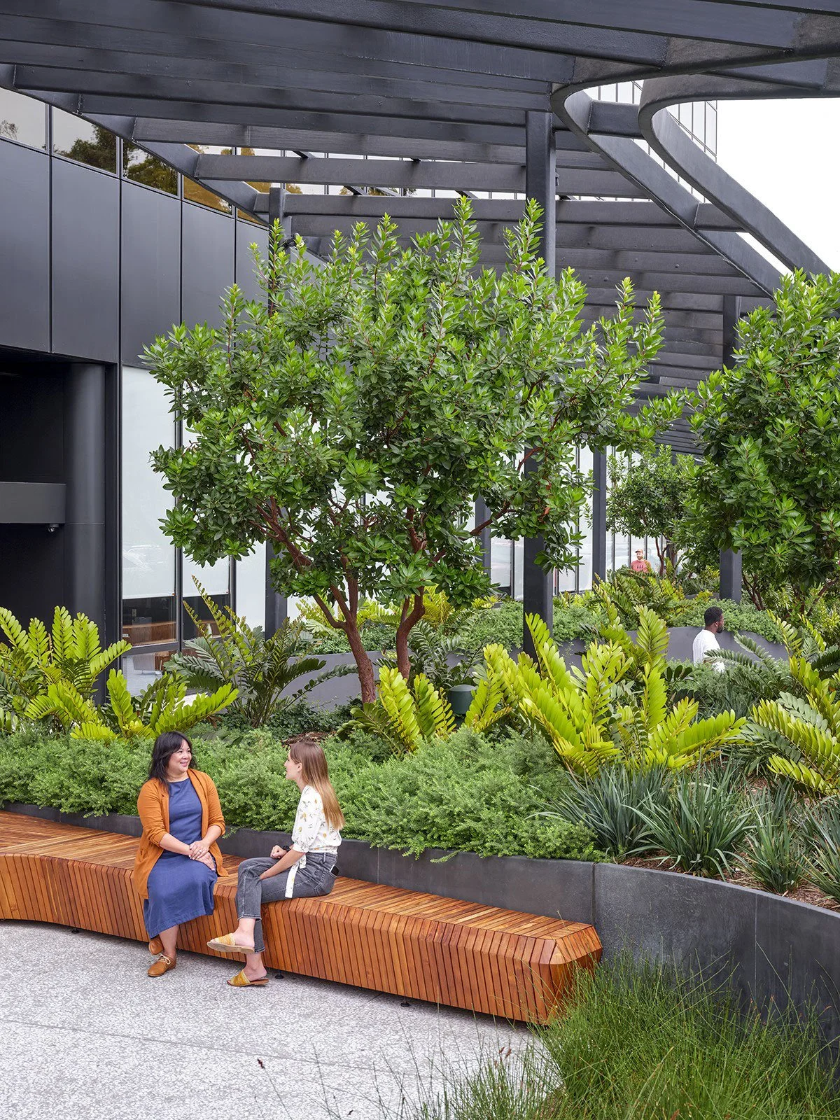 Two women sitting on a curved wooden bench and talking, surrounded by lush green plants and trees in an outdoor urban garden or park setting with modern building in the background.