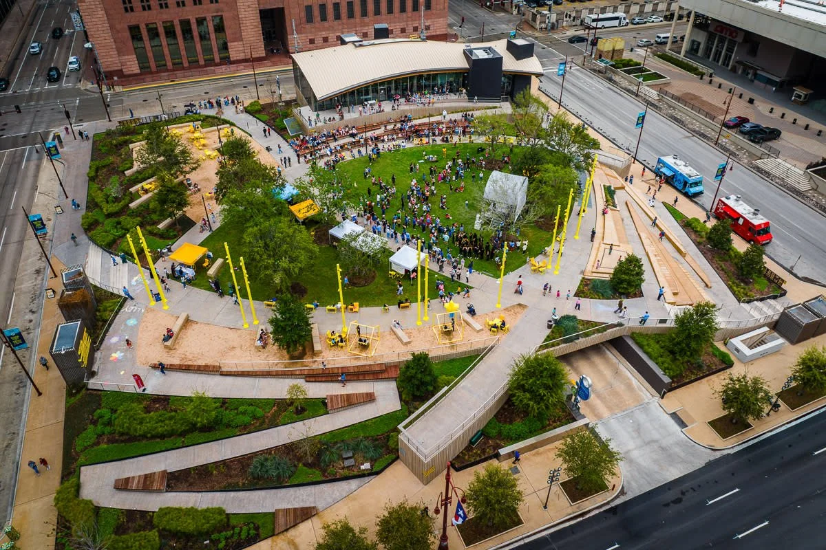 An aerial view of a lively urban park with green grass, trees, and a stage where a large crowd of people is gathered. Surrounding the park are sidewalks, benches, and pathways, with buildings and streets in the background. There are also colorful pol