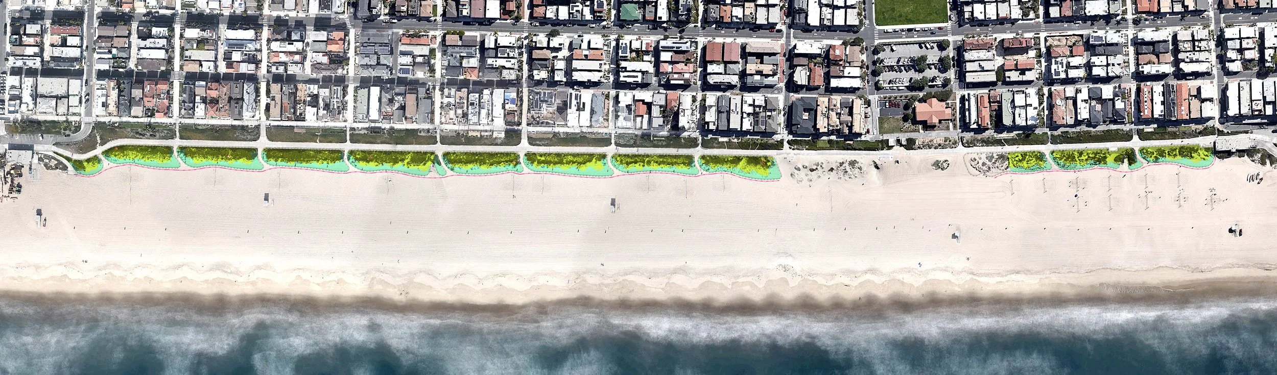 Aerial view of a beachfront with a row of houses behind a green walkway lined with trees along the sandy beach and ocean.