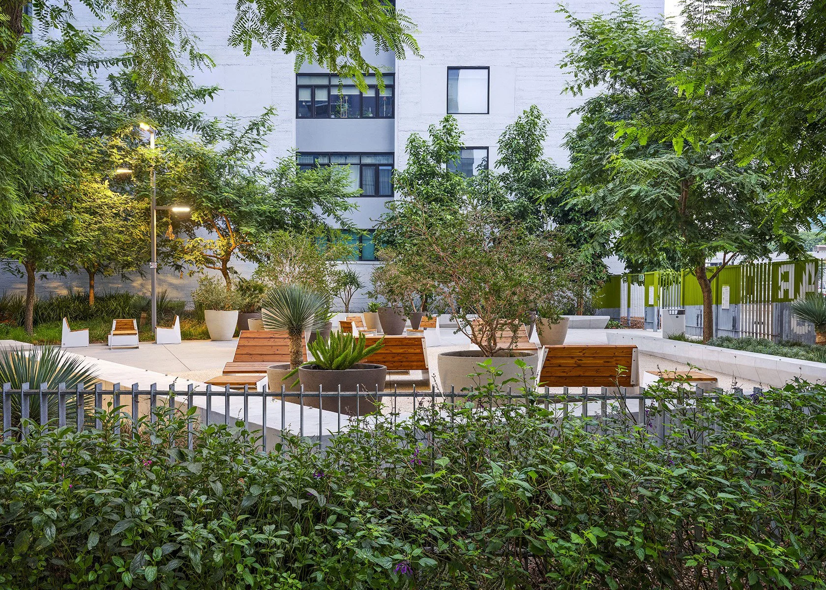 Urban park with wooden benches, large potted plants, and trees, surrounded by a building and a fence, illuminated by streetlights at dusk.