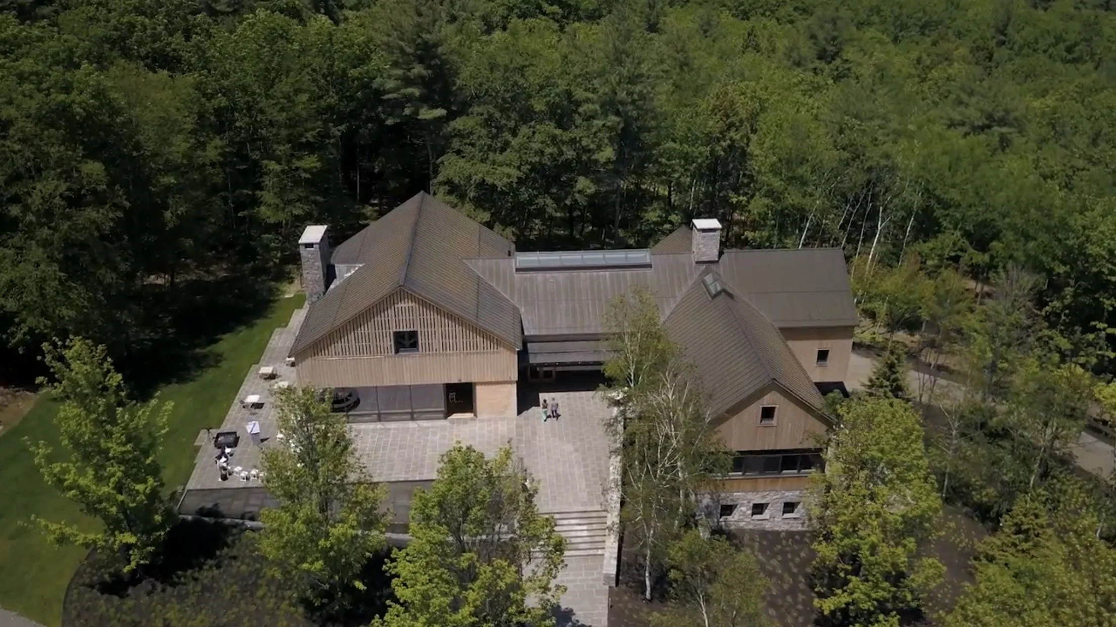 Aerial view of a large house surrounded by trees, with a paved patio area and a lawn, in a wooded area.