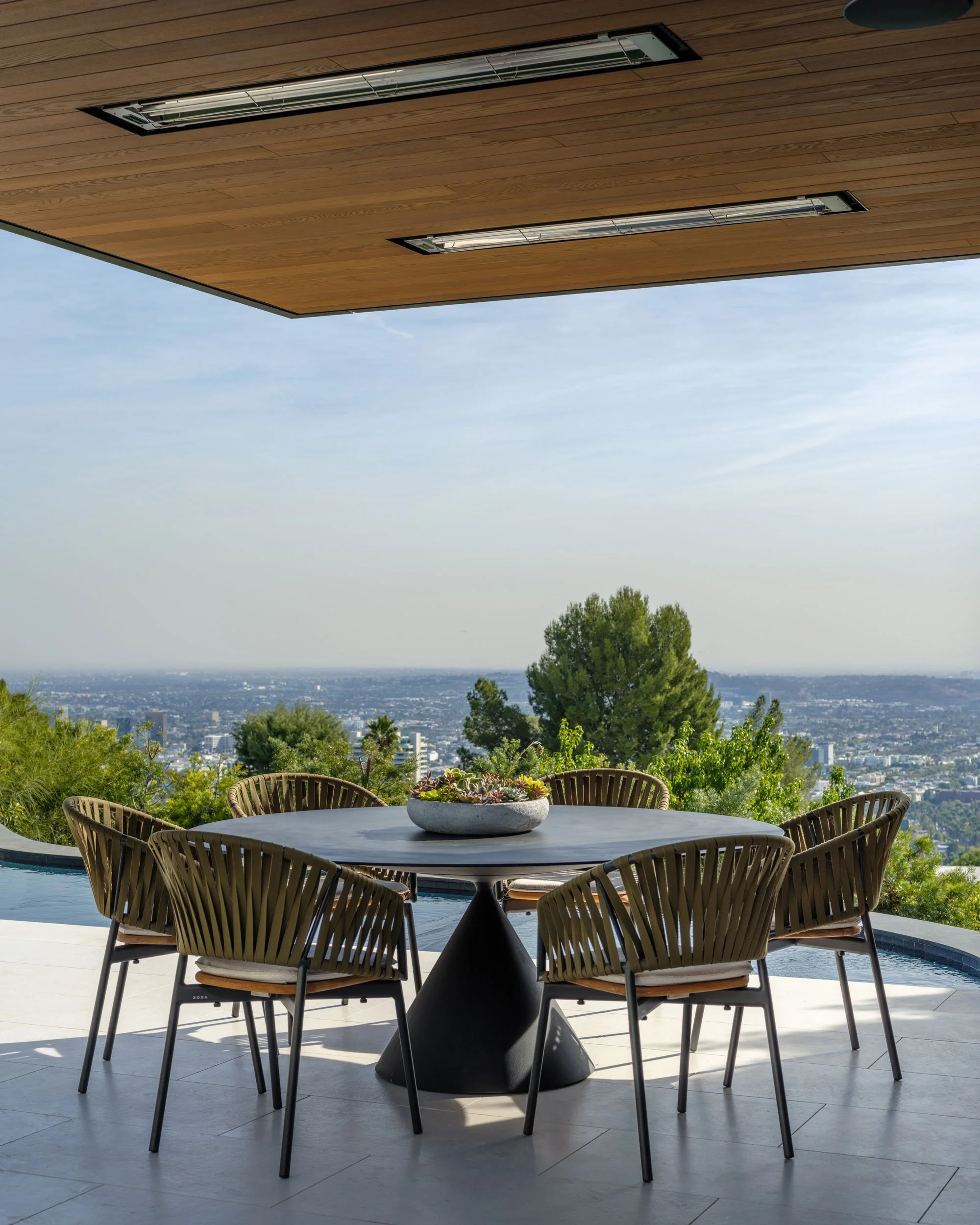 Outdoor patio with a round table and six wicker chairs, a decorative planter with succulents, overlooking a city skyline and trees, with a wooden ceiling exposed from a new garden pavilion.