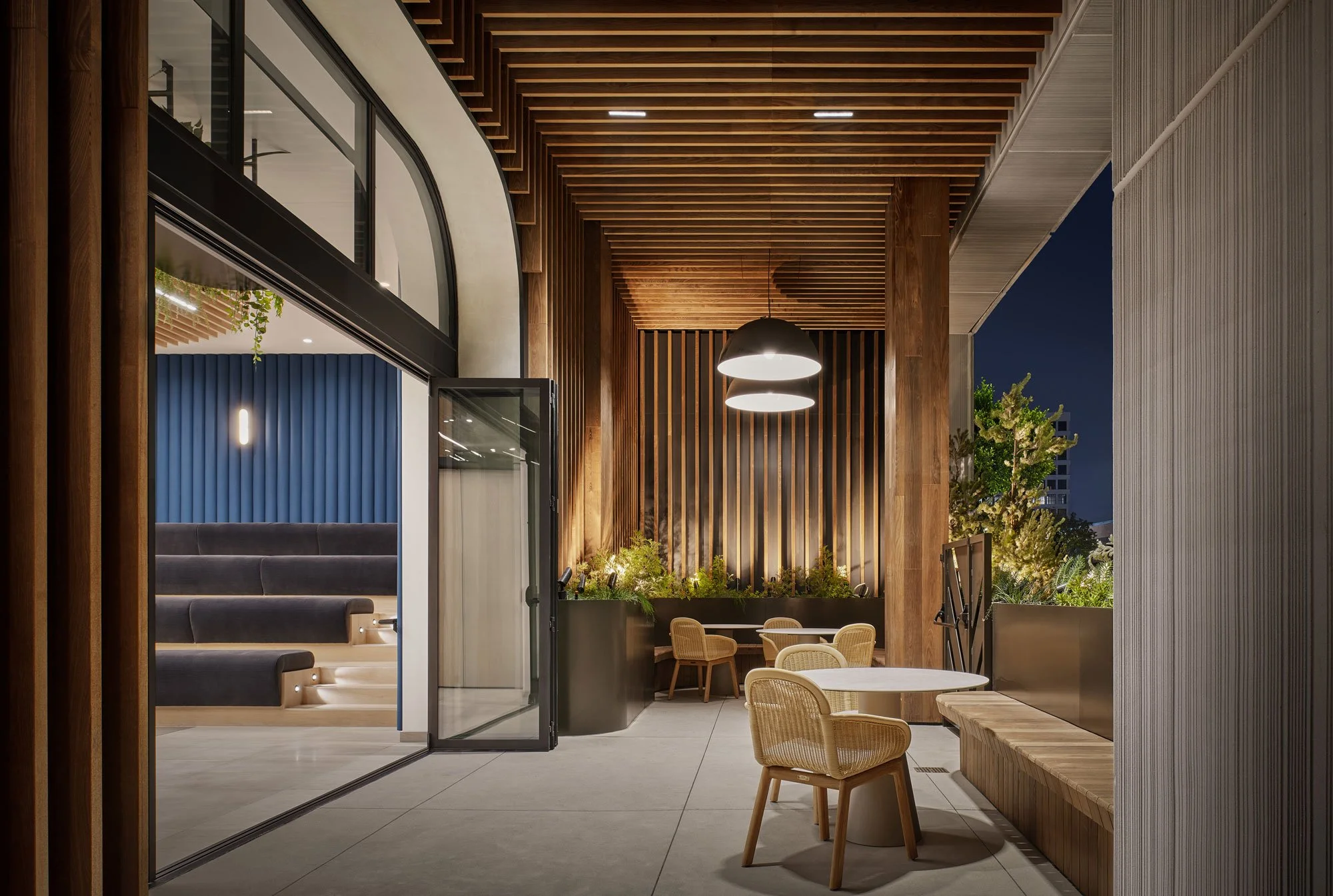 Modern indoor-outdoor seating area at night with wooden slats on ceiling, hanging light fixtures, a plant wall, chairs, and a view of the dark sky outside.