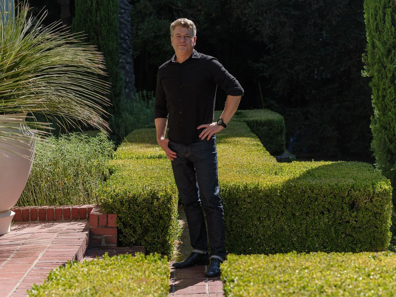 A portrait of Peter Emerson, Owner and Creative Director of Emerson Collab, standing outdoors in a garden with neatly trimmed bushes and trees, wearing a black shirt and dark jeans, facing the camera with one hand on his hip.