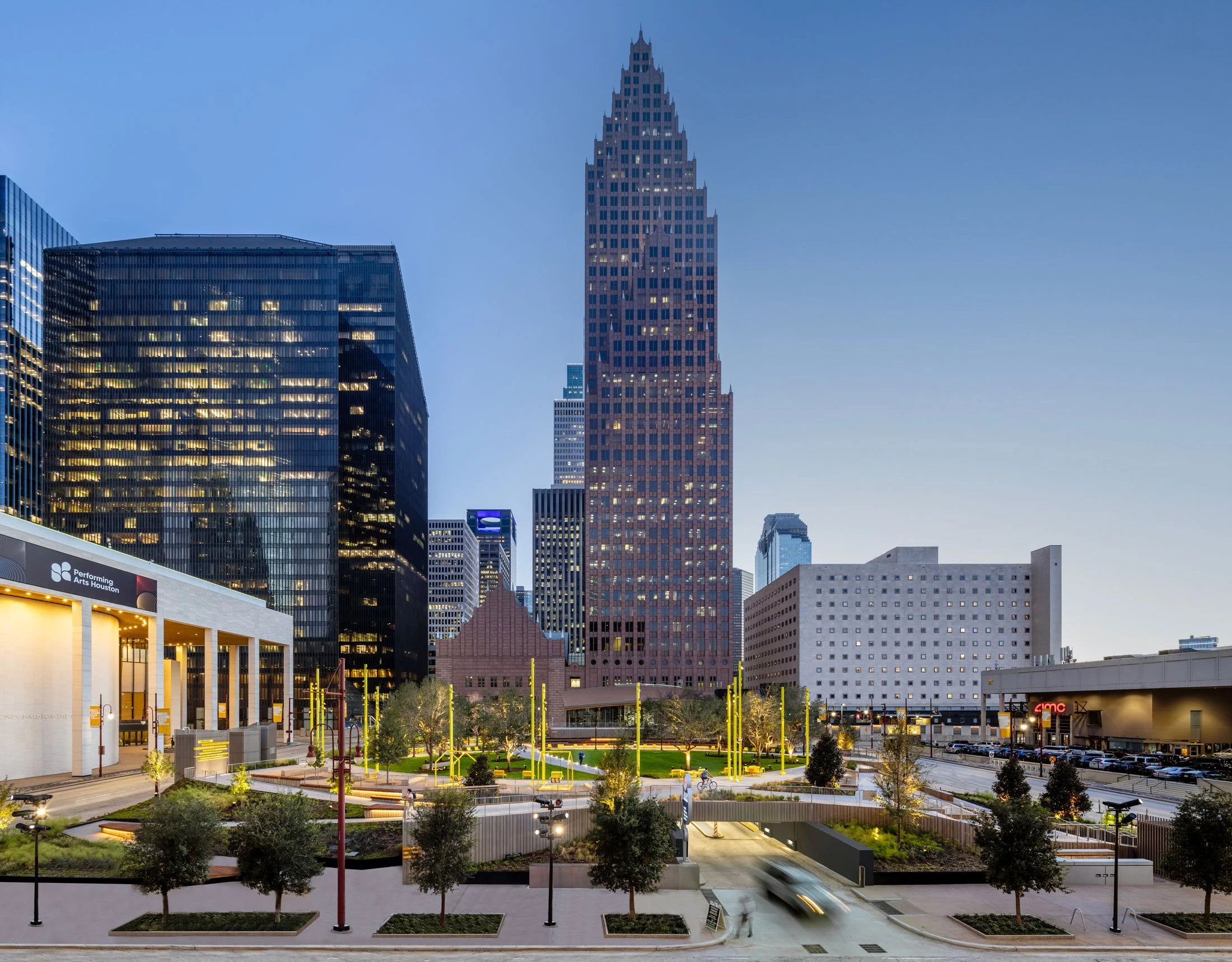 A cityscape of Houston, Texas, featuring the 1000 Louisiana Street building in the center, with a park in the foreground and modern glass office buildings on the left. The sky is clear with a slight gradient from daylight to early evening.