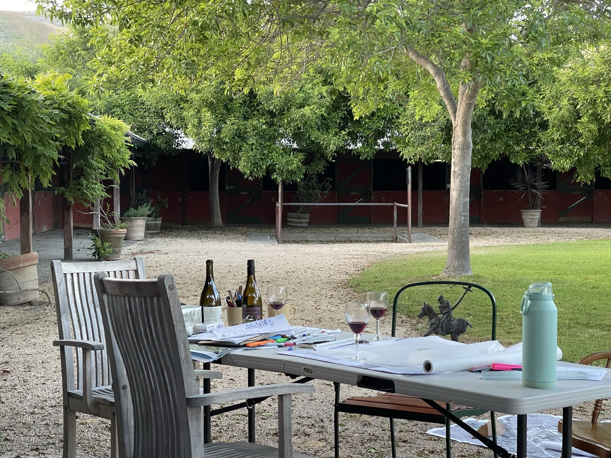 Outdoor patio area with a table set for dining, featuring wine glasses, bottles, papers, and pens. Surrounding the table are wooden chairs, and in the background, there are trees and potted plants, with a building and a fence visible behind the green