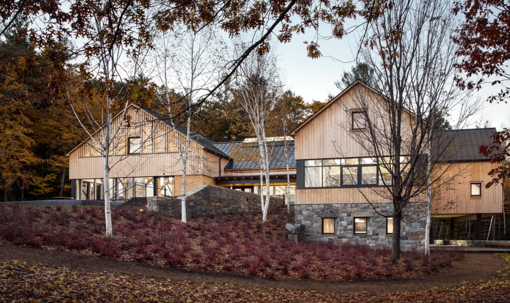 A modern, multi-story house with a wooden exterior, large windows, and a stone foundation, set among trees with brown and red autumn leaves.