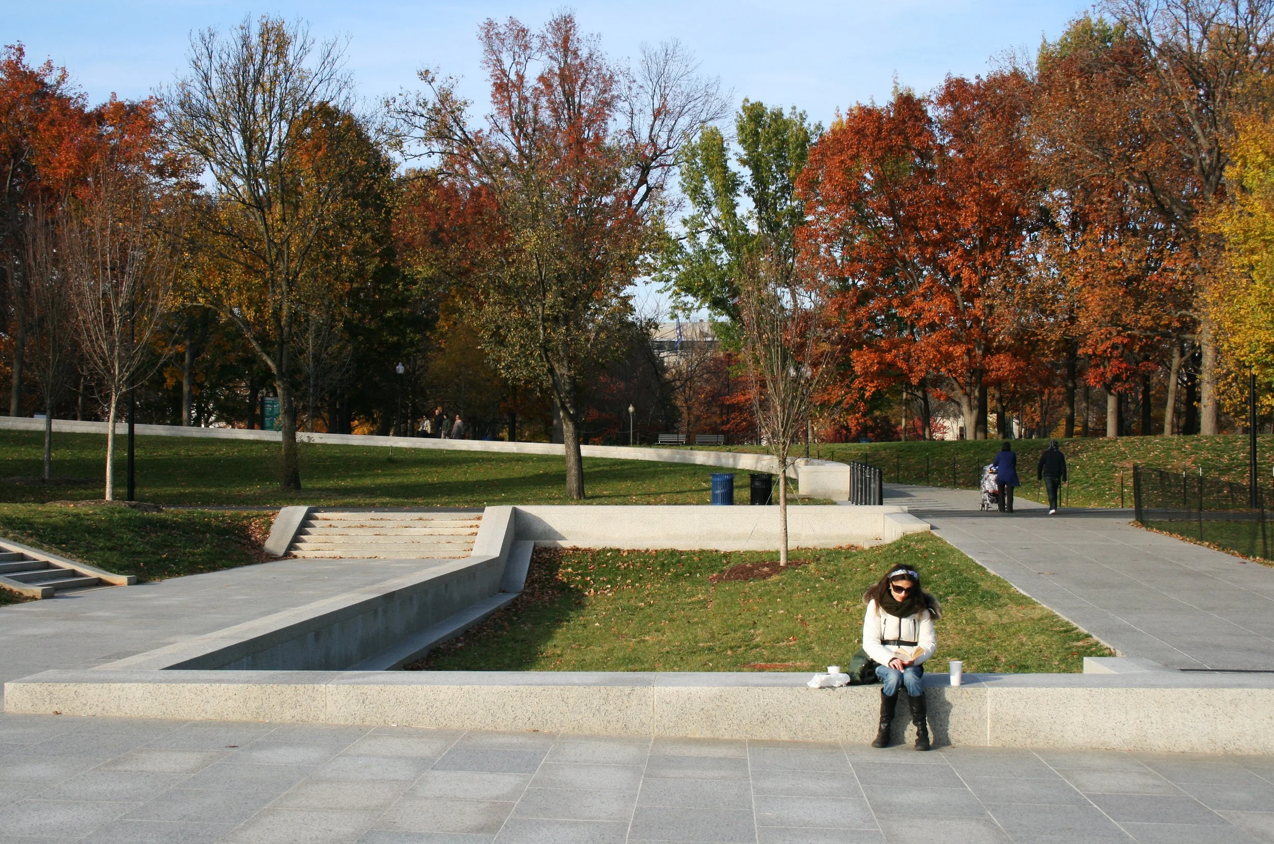 A woman sitting in a rectangular concrete sunken seat in a park, wearing sunglasses, a white coat, and black boots, with a coffee cup on her lap. In the background, there are trees with fall foliage, a pathway with a couple walking with a stroller, a
