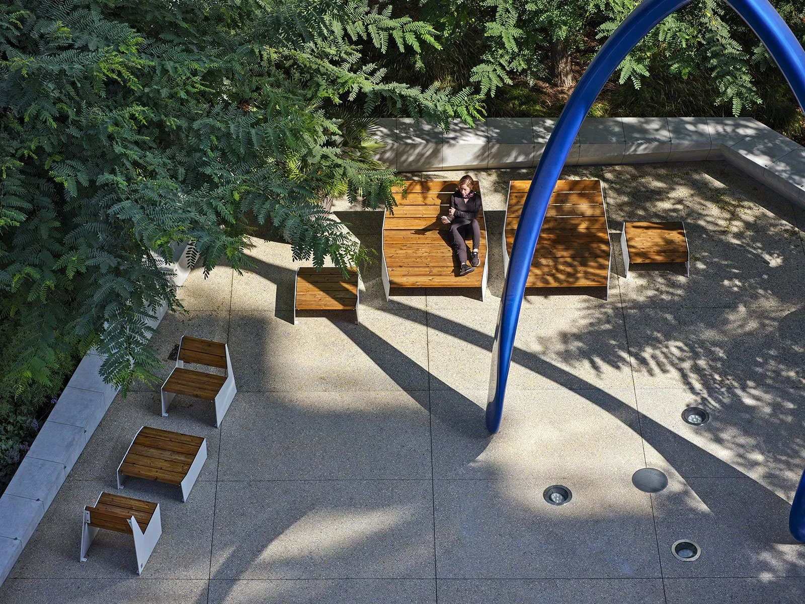 A woman is lying on a wooden lounge chair in a sunny outdoor space, surrounded by trees and shade, with several more wooden benches or lounge chairs nearby on a paved ground under a sculpture by Desire Obtain Cherish entitled 'Lucky Break.'