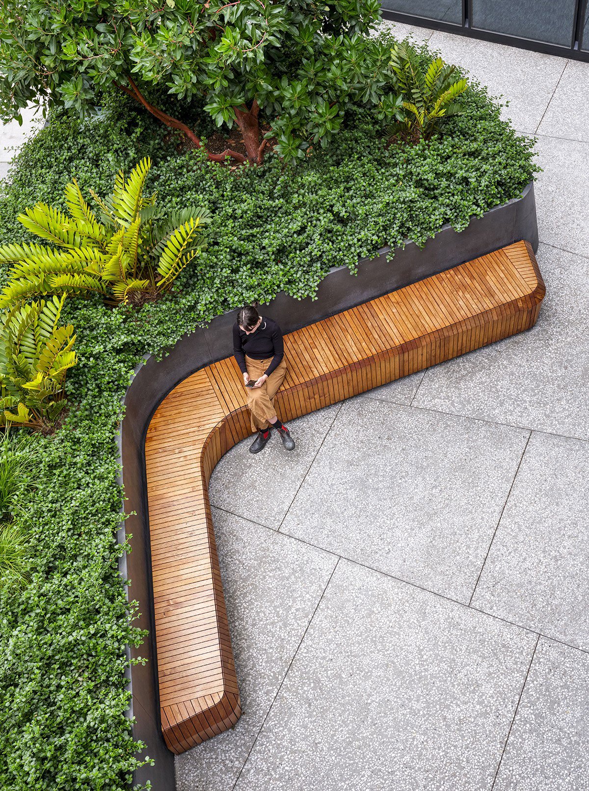 A person sitting on a curved wooden bench outdoors, surrounded by green bushes and plants, with exposed aggregate paving and a building wall in the background.