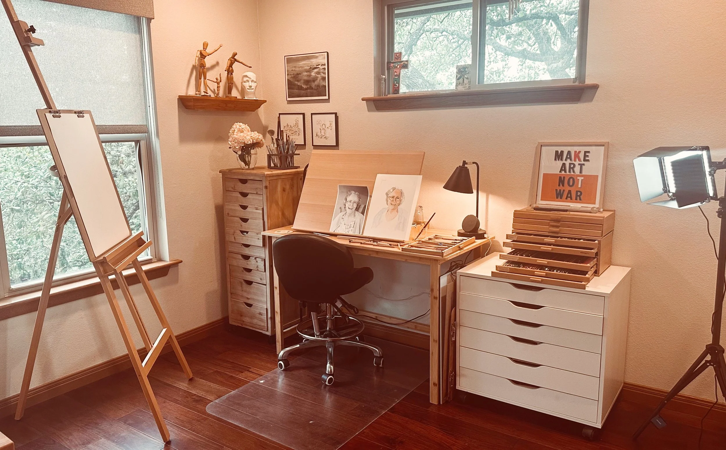 A cozy art studio with wooden flooring, featuring a wooden easel near a window, a desk with art supplies, paintings of older women on the desk, framed artwork on the wall, a sign that reads 'Make Art Not War,' and various art-themed decorations. Daylight filters through the windows, illuminating the space.