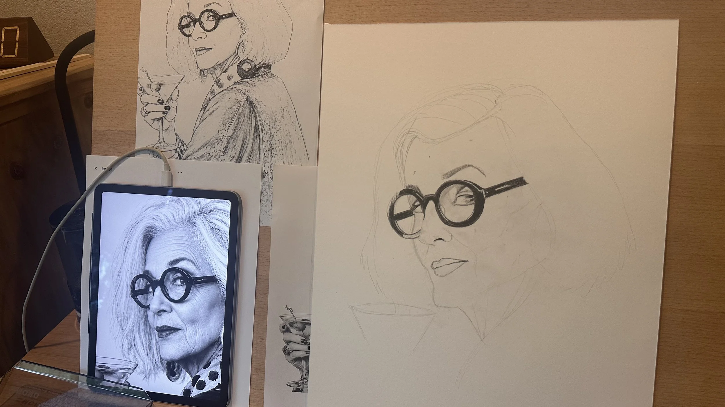 Process of creating a portrait, showing a printed black-and-white photo of an elderly woman with glasses and long hair, a sketched outline of the same woman's face, and a partially completed artistic drawing of her.