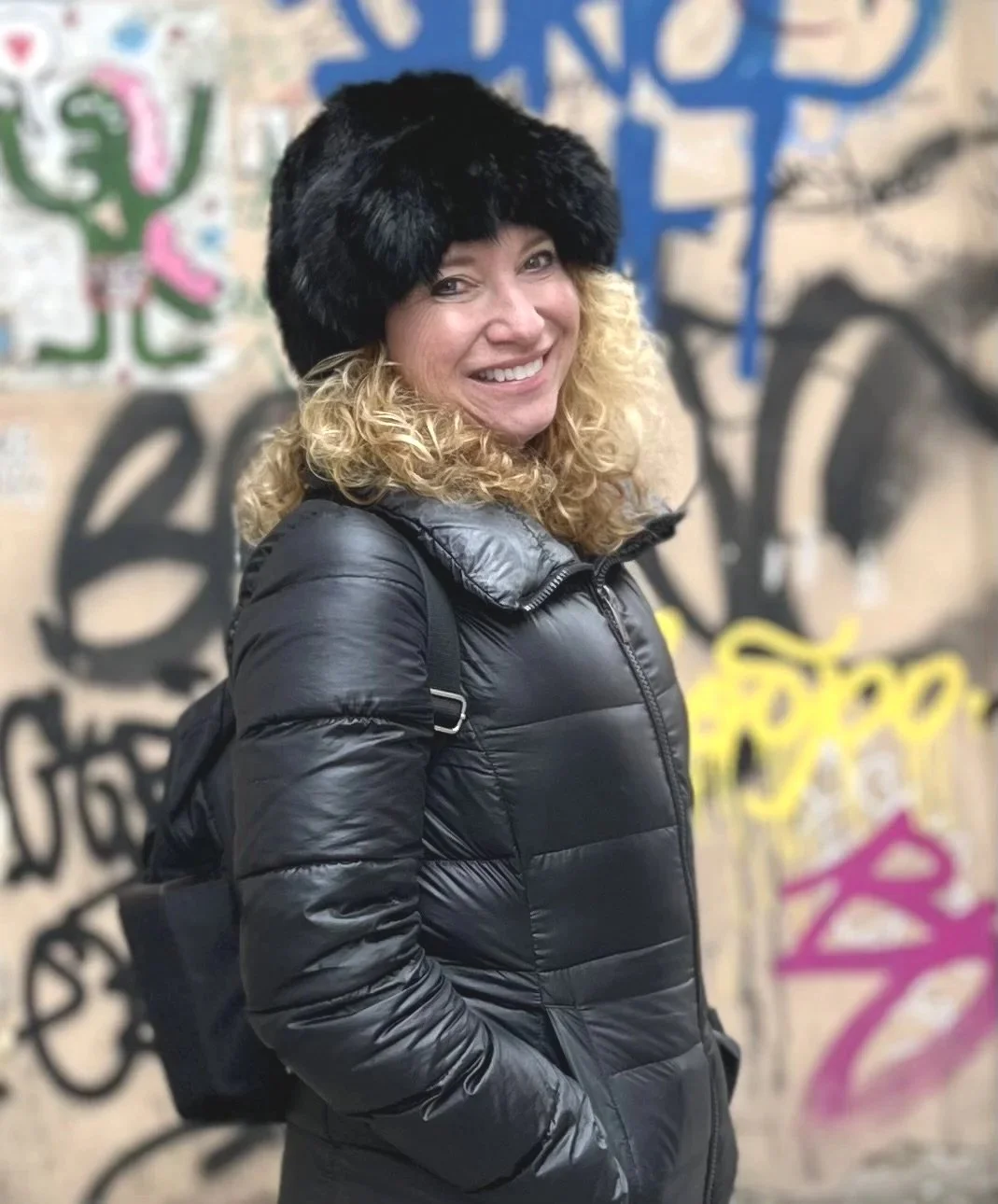 Woman wearing a black fur hat and black puffy jacket smiling in front of a graffiti-covered wall.