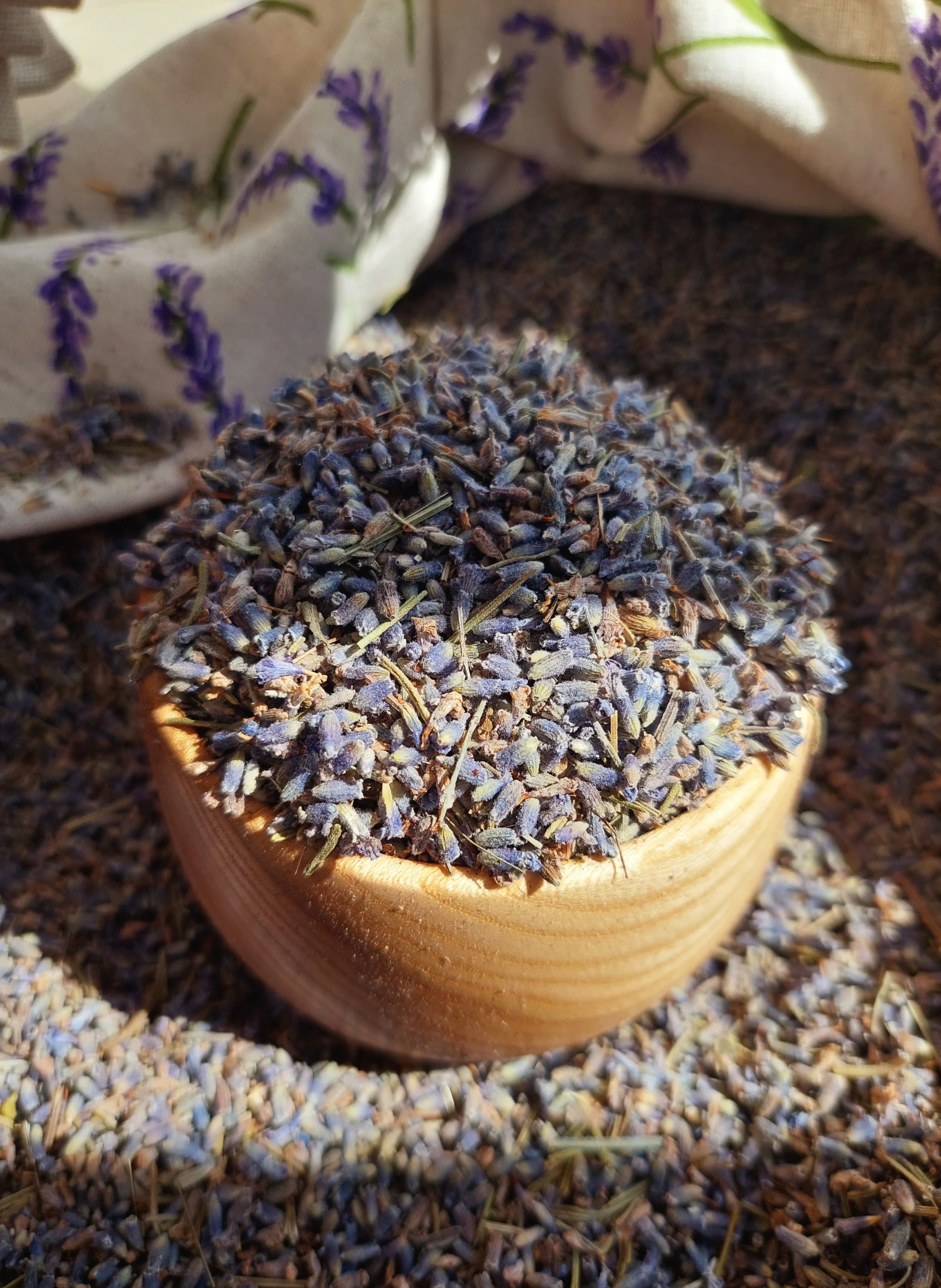 Close-up of dried lavender flowers in a wooden bowl, with a linen cloth featuring lavender flowers in the background, on a surface covered with lavender buds.