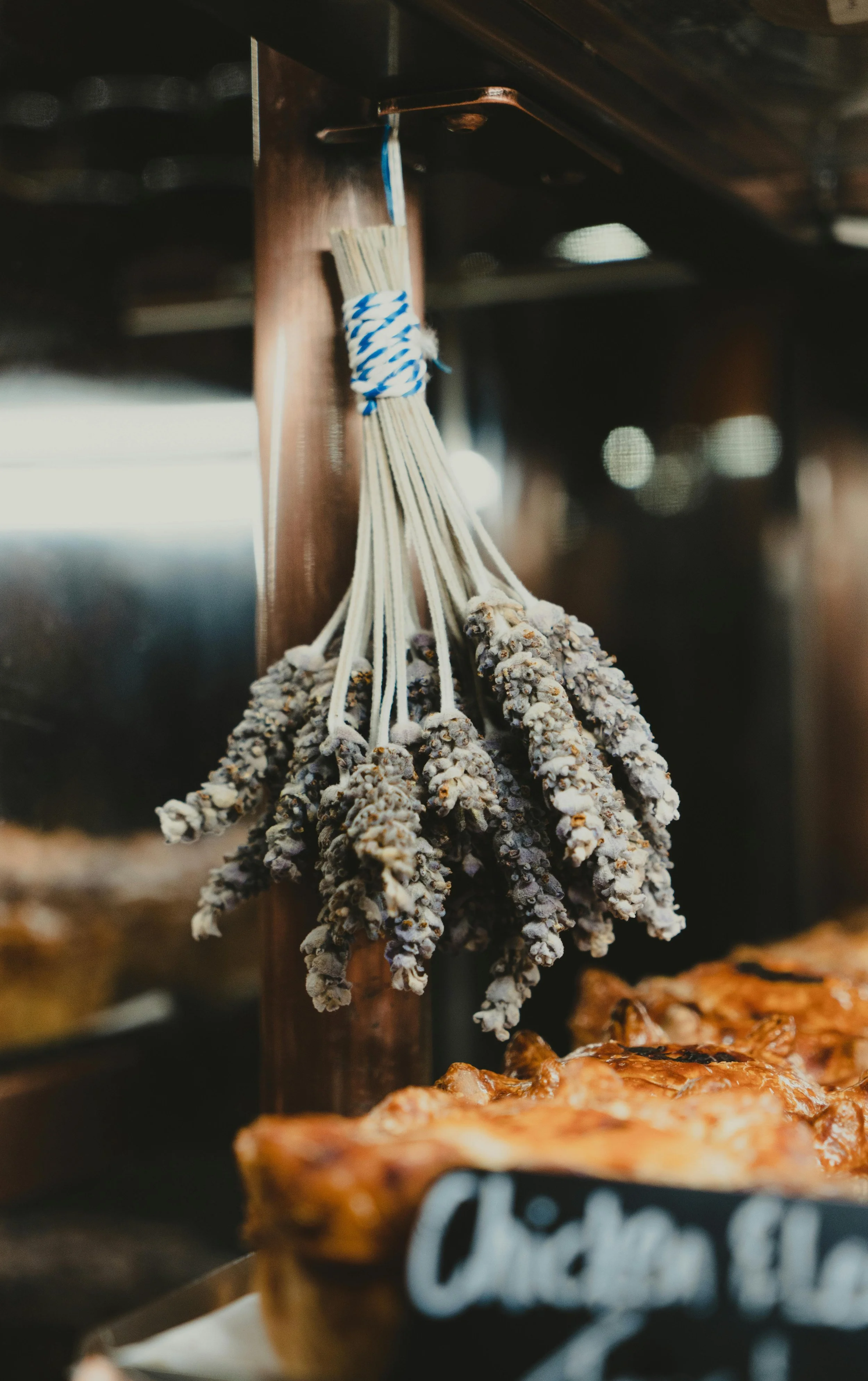 Bunches of dried lavender hanging upside down inside a bakery or kitchen, with a blurred background of baked goods.