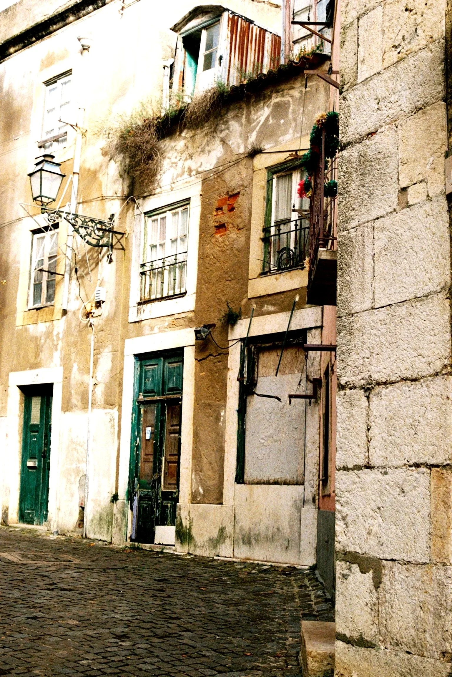 An old, weathered building with multiple windows and doors, some with shutters, on a cobblestone street. The building has a mix of plaster and exposed brick, with some plants on a small balcony and a street lamp attached to the wall.