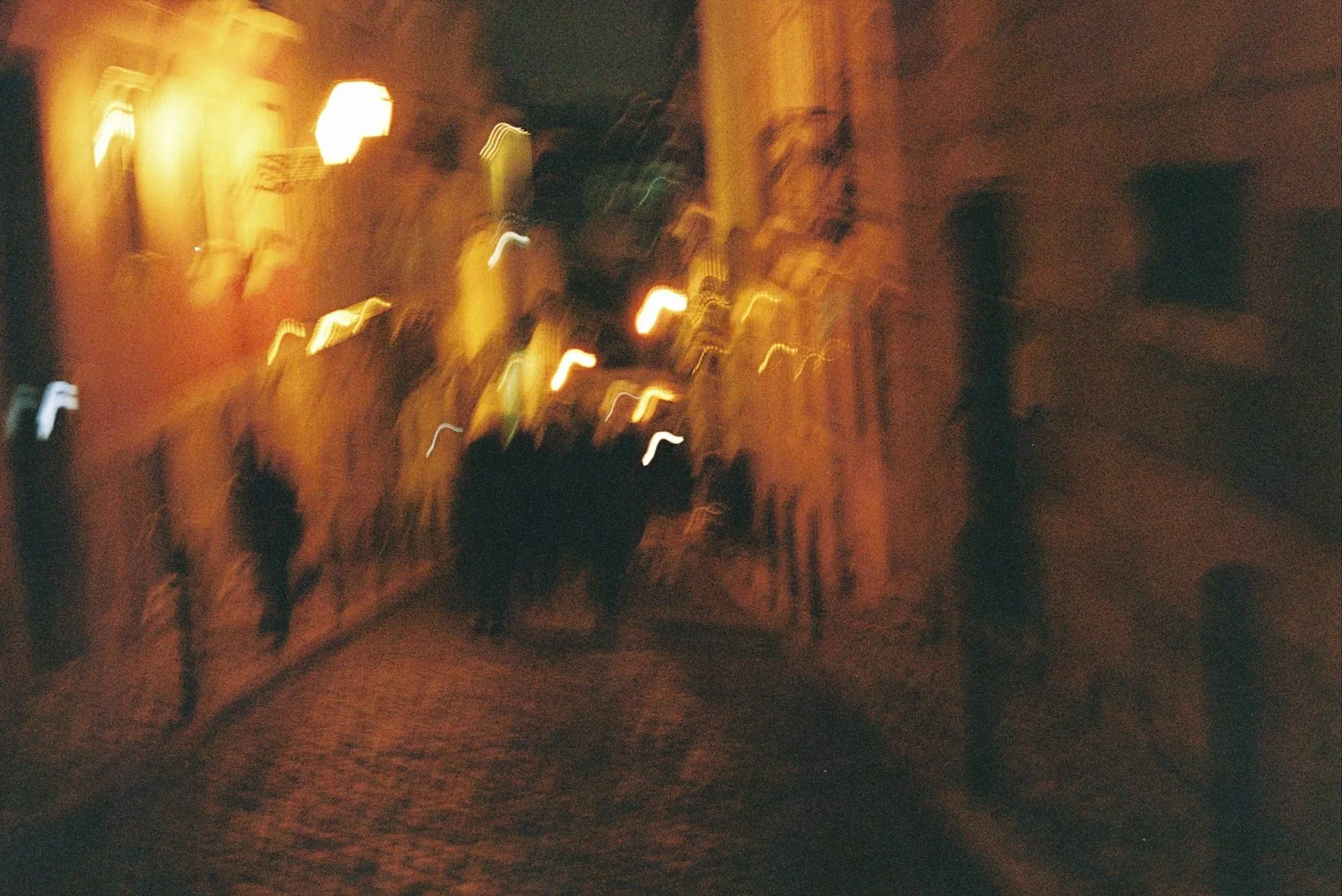 Nighttime street scene with blurred, glowing streetlights and silhouettes of people walking on the sidewalk.