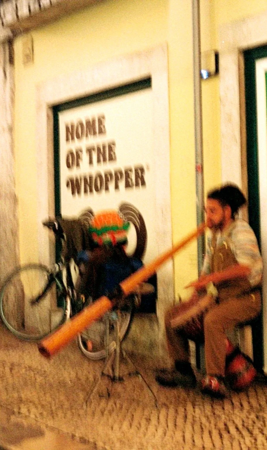 A person sitting on a stool outside a building, playing a didgeridoo, with a coffee bicycle and a sign that reads "Home of the Whopper" in the background.