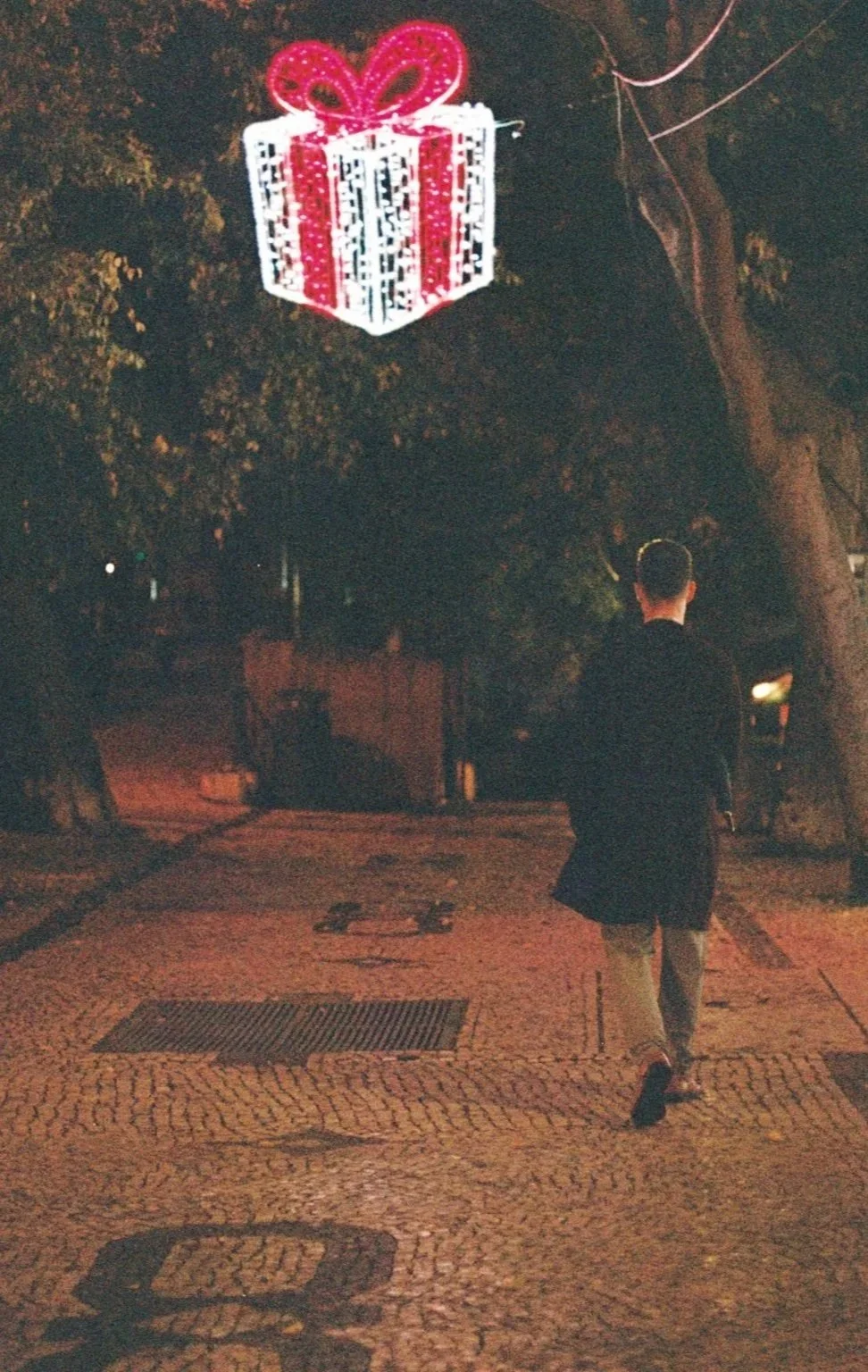 A man walking on a cobblestone street at night with a large illuminated gift box decoration hanging from a tree above him.