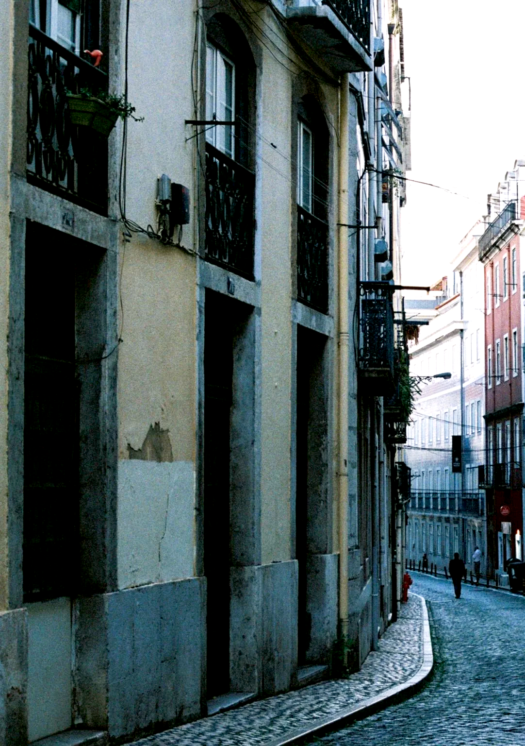 Narrow street with cobblestone pavement, old buildings with balconies, some with plants, and a few pedestrians in the distance.