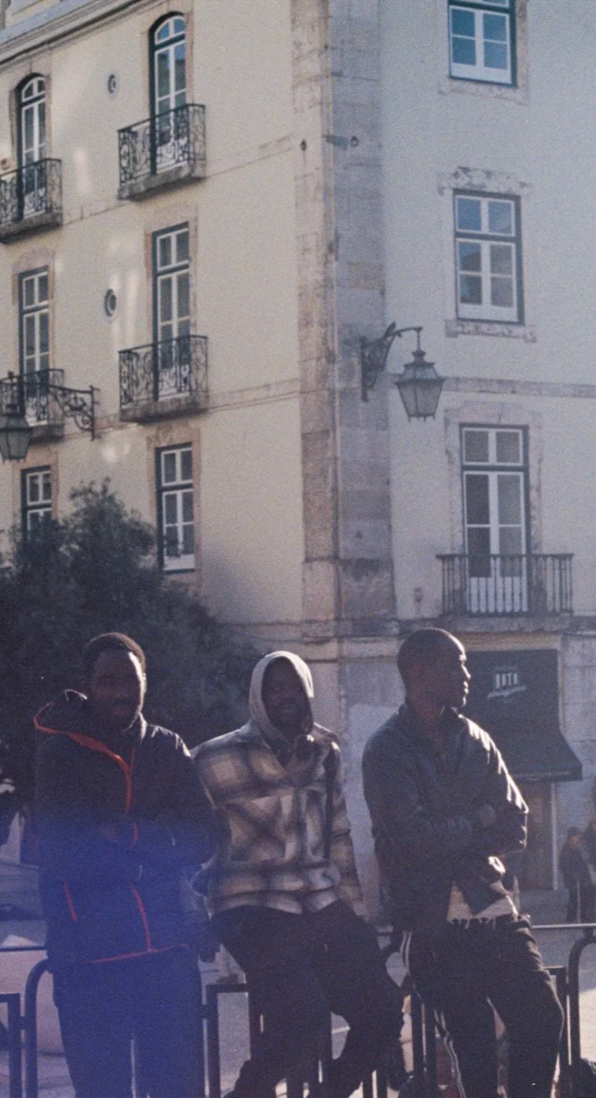 Three young men standing outdoors in front of a multi-story building with European-style architecture, featuring windows with balconies and lamps, during daytime.